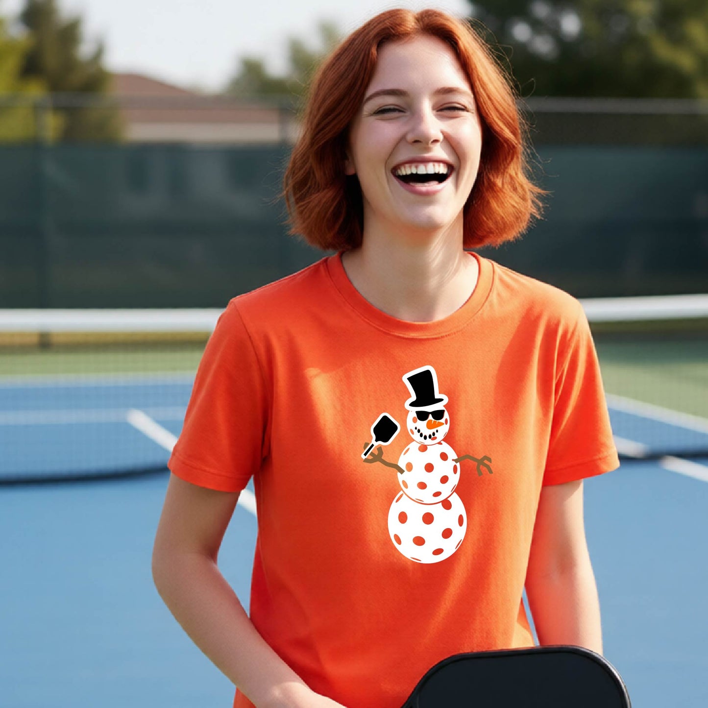 A woman with red hair laughing on a pickleball  court, wearing a bright orange t-shirt with a cartoon snowman pickleball graphic.
