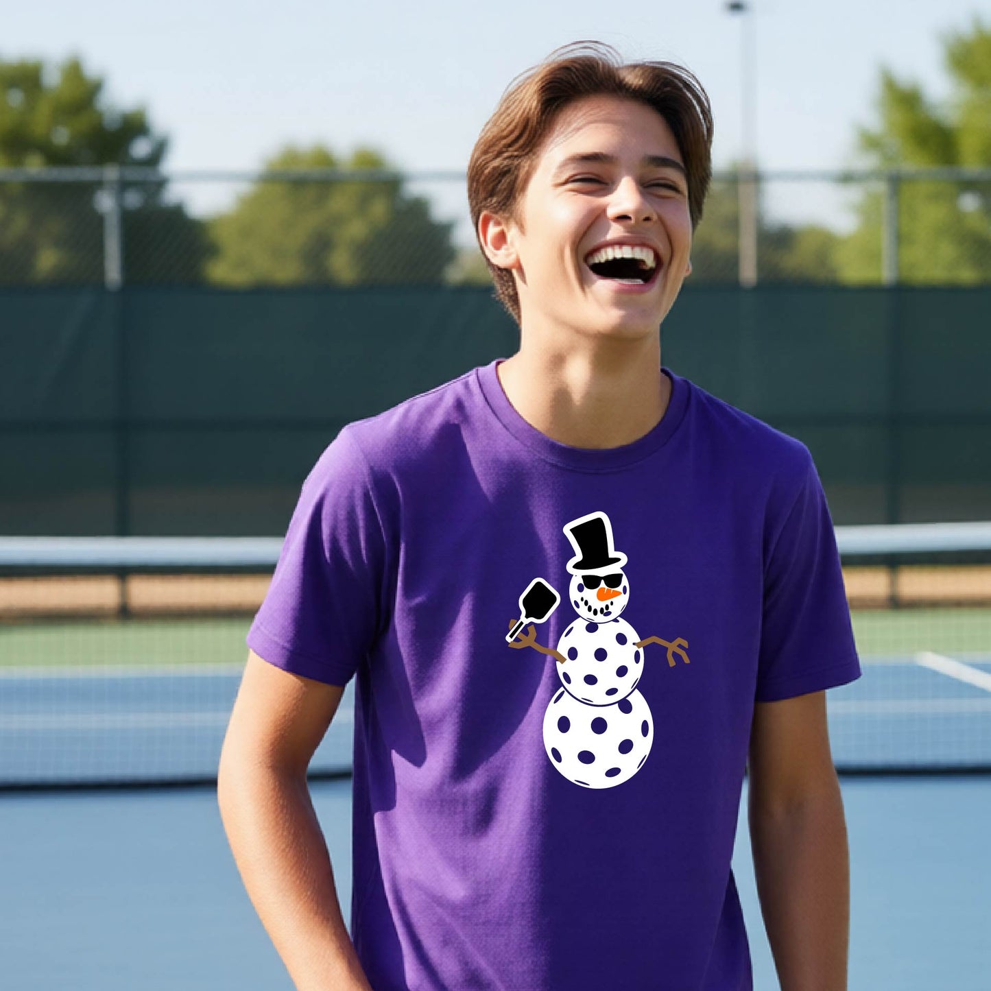 A man laughing on an outdoor pickleball court wearing a purple t-shirt with a cartoon snowman pickleball graphic.