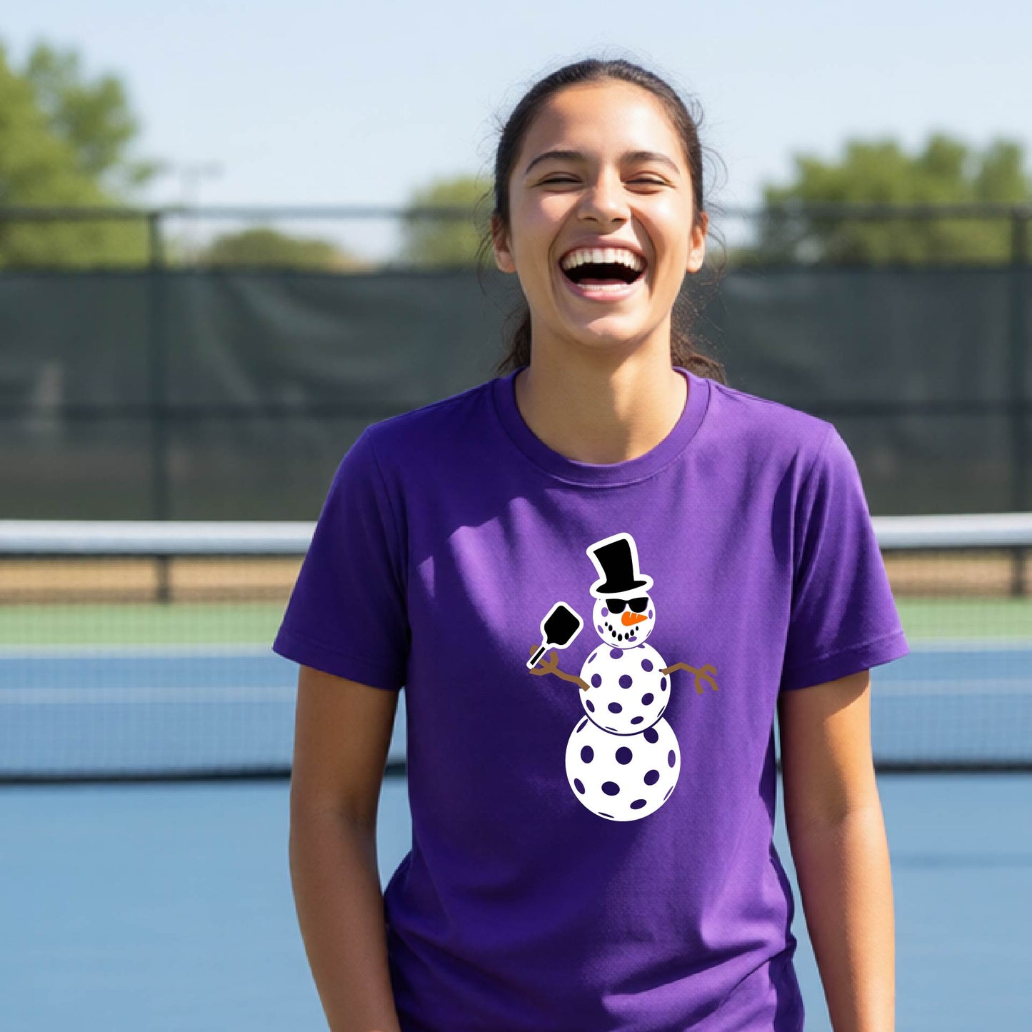 A woman laughing on an outdoor pickleball  court wearing a purple t-shirt with a cartoon snowman pickleball graphic.