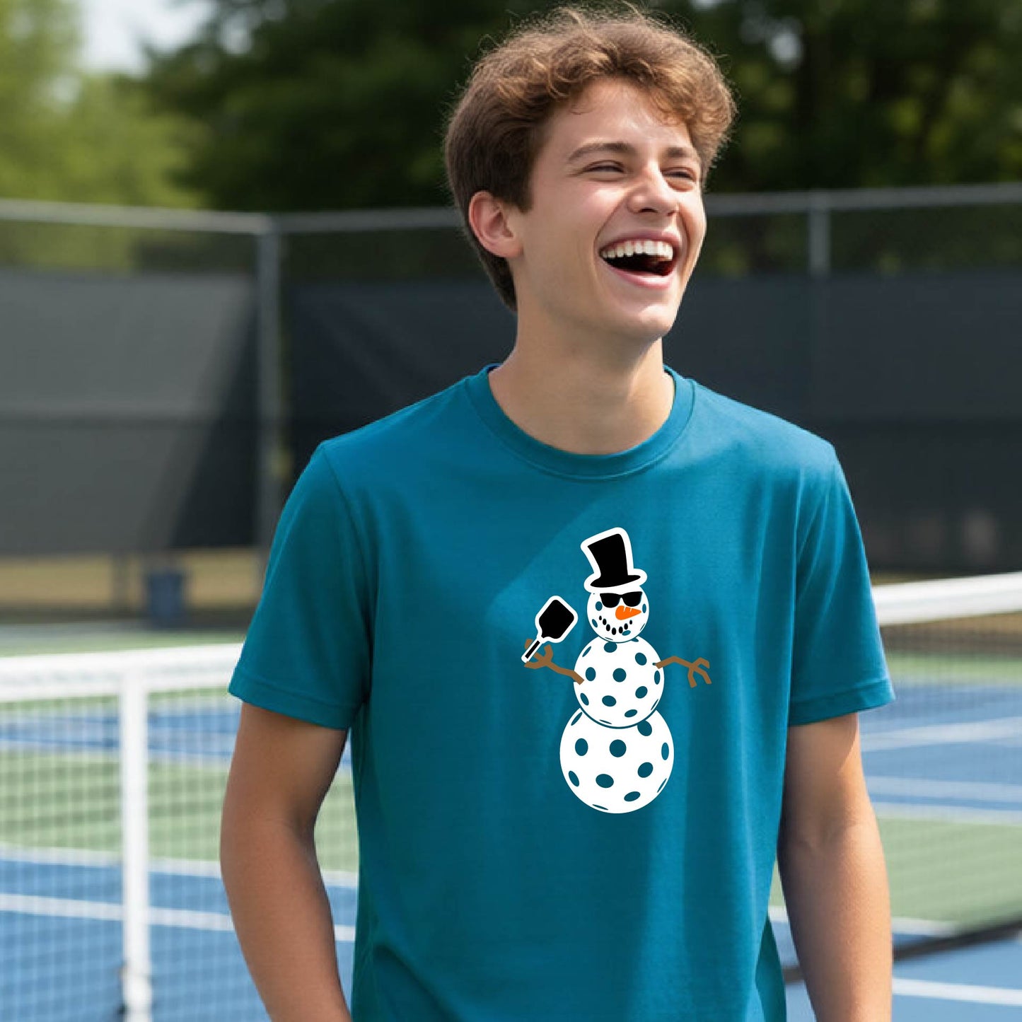 A young man laughing on an outdoor pickleball  court wearing a teal t-shirt with a cartoon snowman pickleball graphic on the front.