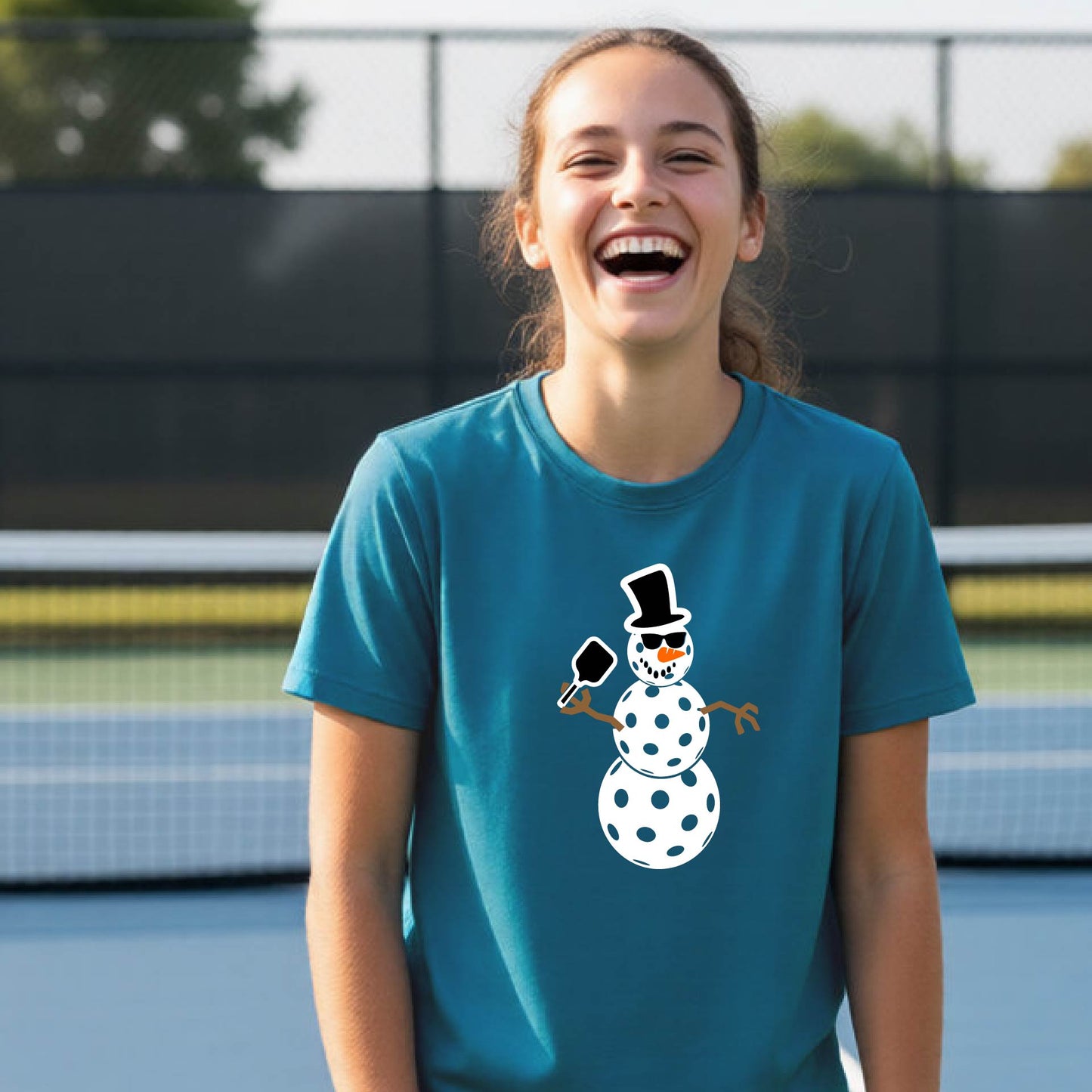 A young woman laughing on an outdoor pickleball  court, wearing a teal blue t-shirt with a cartoon snowman pickleball graphic on the front.