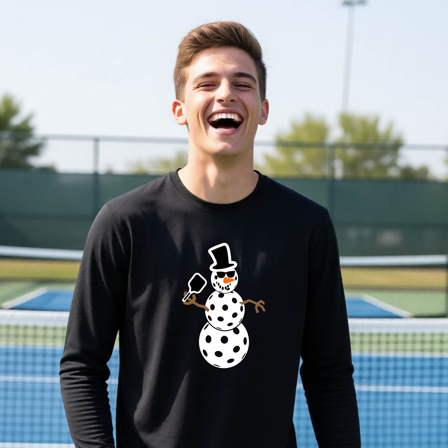 A smiling young person stands on a pickleball court wearing a black long-sleeve shirt with a white and pink polka-dot pickleball snowman graphic on the front.