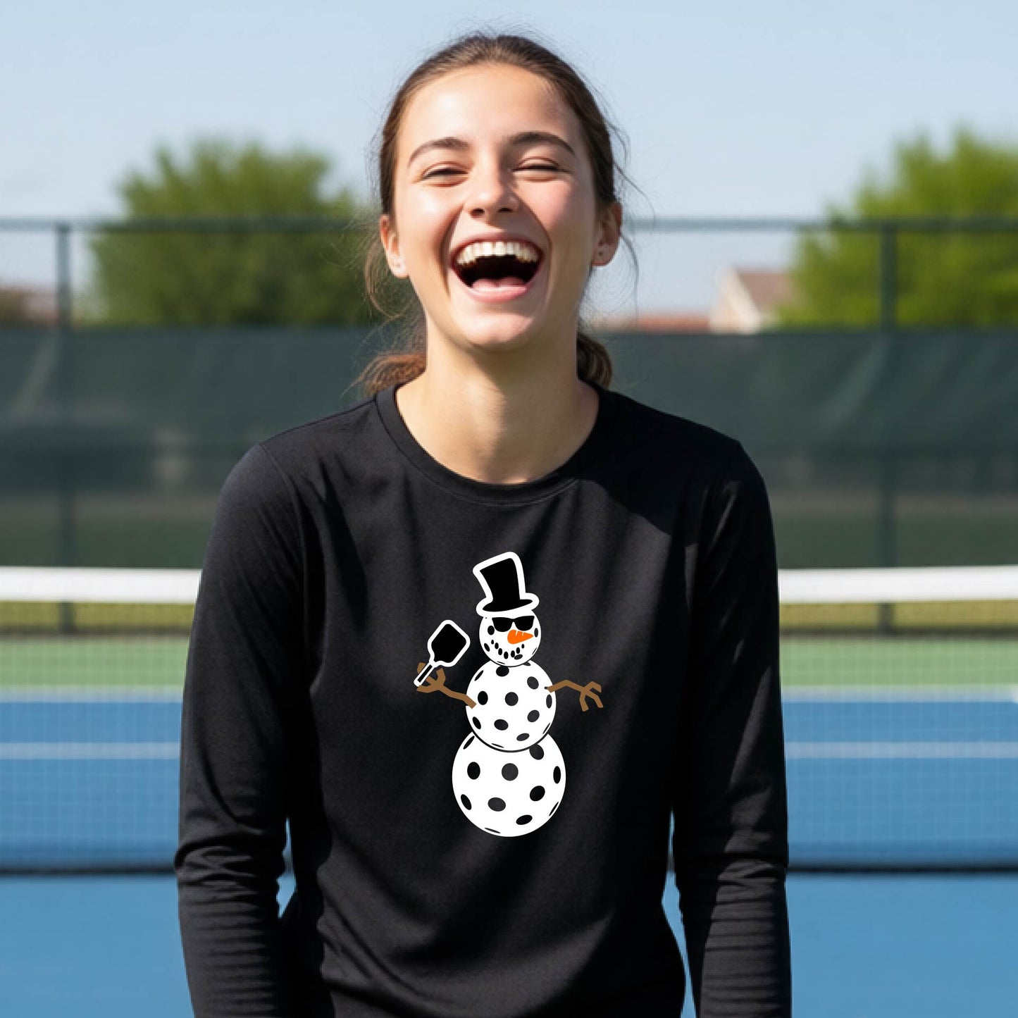 A smiling young person stands on a pickleball court wearing a black long-sleeve shirt with a white and pink polka-dot pickleball snowman graphic on the front.