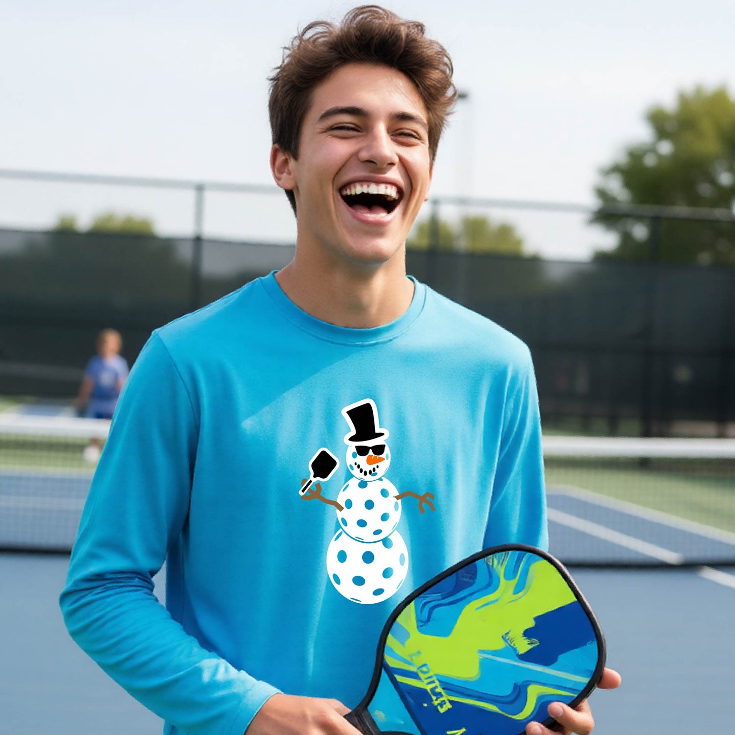 A smiling young person stands on a pickleball court wearing a bright blue long-sleeve shirt with a white and pink polka-dot pickleball snowman graphic on the front.