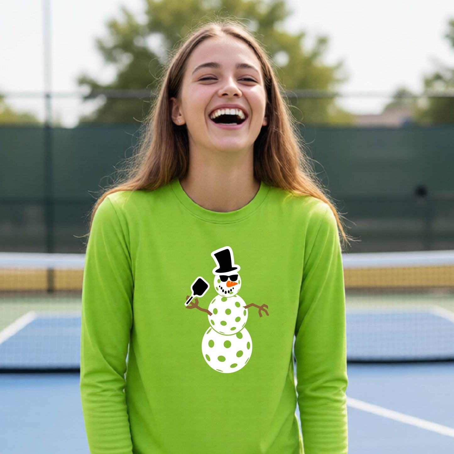 A smiling young person stands on a pickleball court wearing a bright lime green long-sleeve shirt with a white and pink polka-dot pickleball snowman graphic on the front.