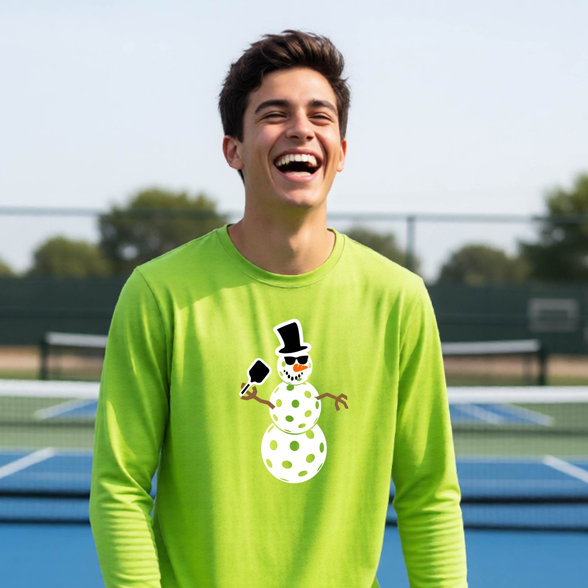 A smiling young person stands on a pickleball court wearing a bright lime green long-sleeve shirt with a white and pink polka-dot pickleball snowman graphic on the front.