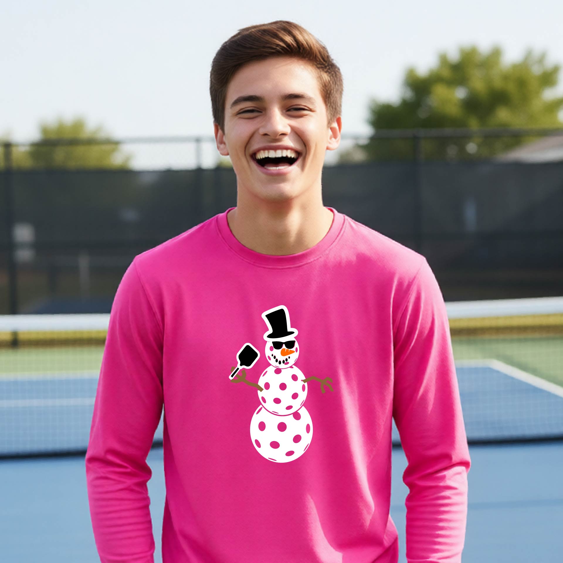 A smiling young person stands on a pickleball court wearing a bright pink long-sleeve shirt with a white and pink polka-dot pickleball snowman graphic on the front.