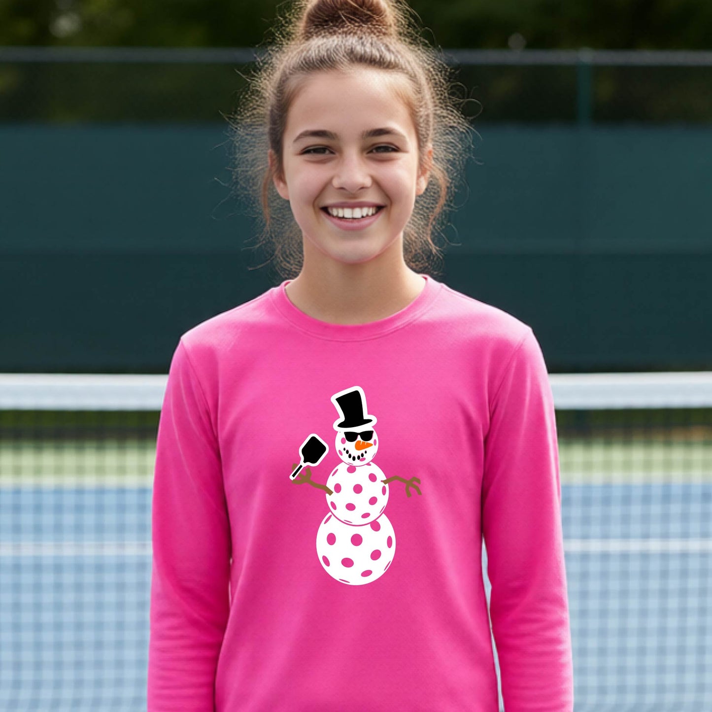 A smiling young person stands on a pickleball court wearing a bright pink long-sleeve shirt with a white and pink polka-dot pickleball snowman graphic on the front.