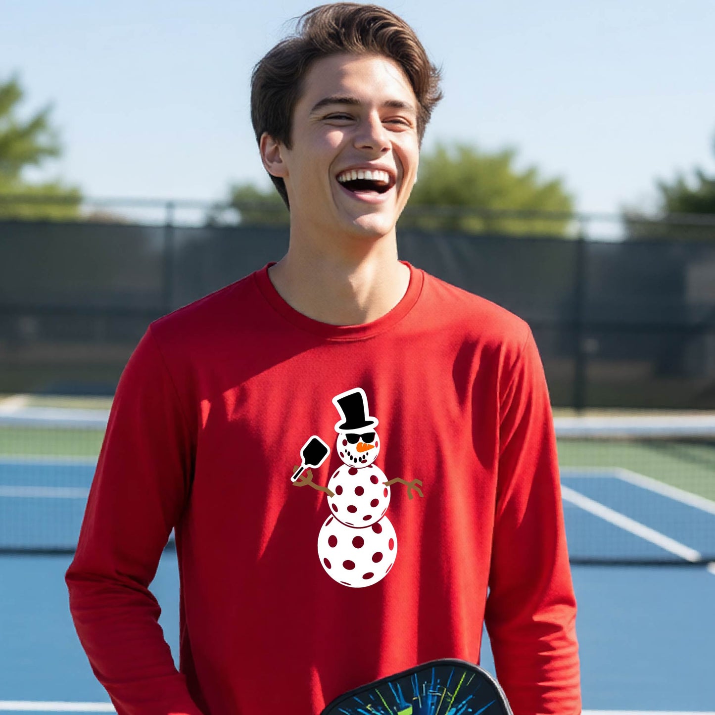 A smiling young person stands on a pickleball court wearing a bright red long-sleeve shirt with a white and pink polka-dot pickleball snowman graphic on the front.