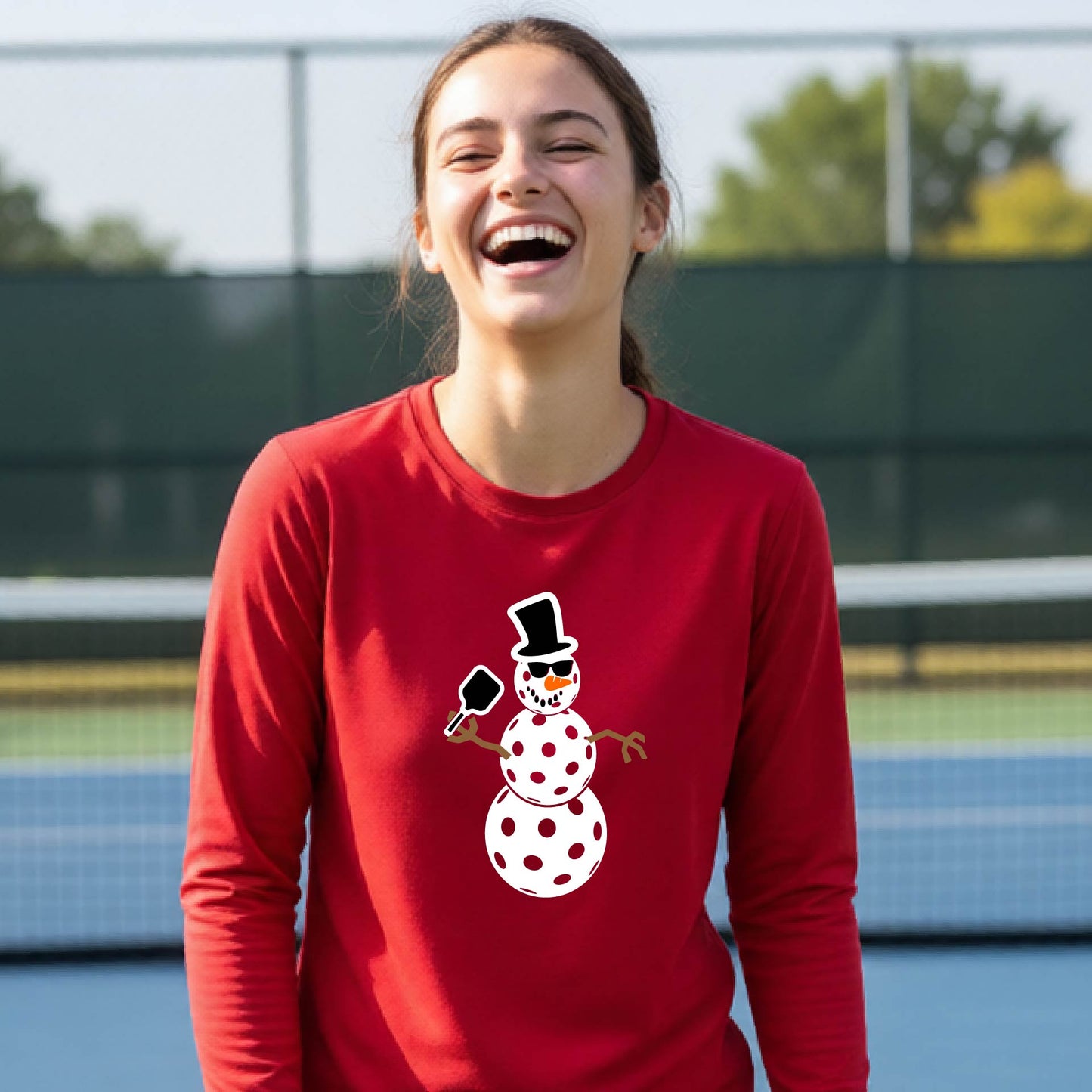 A smiling young person stands on a pickleball court wearing a bright red long-sleeve shirt with a white and pink polka-dot pickleball snowman graphic on the front.