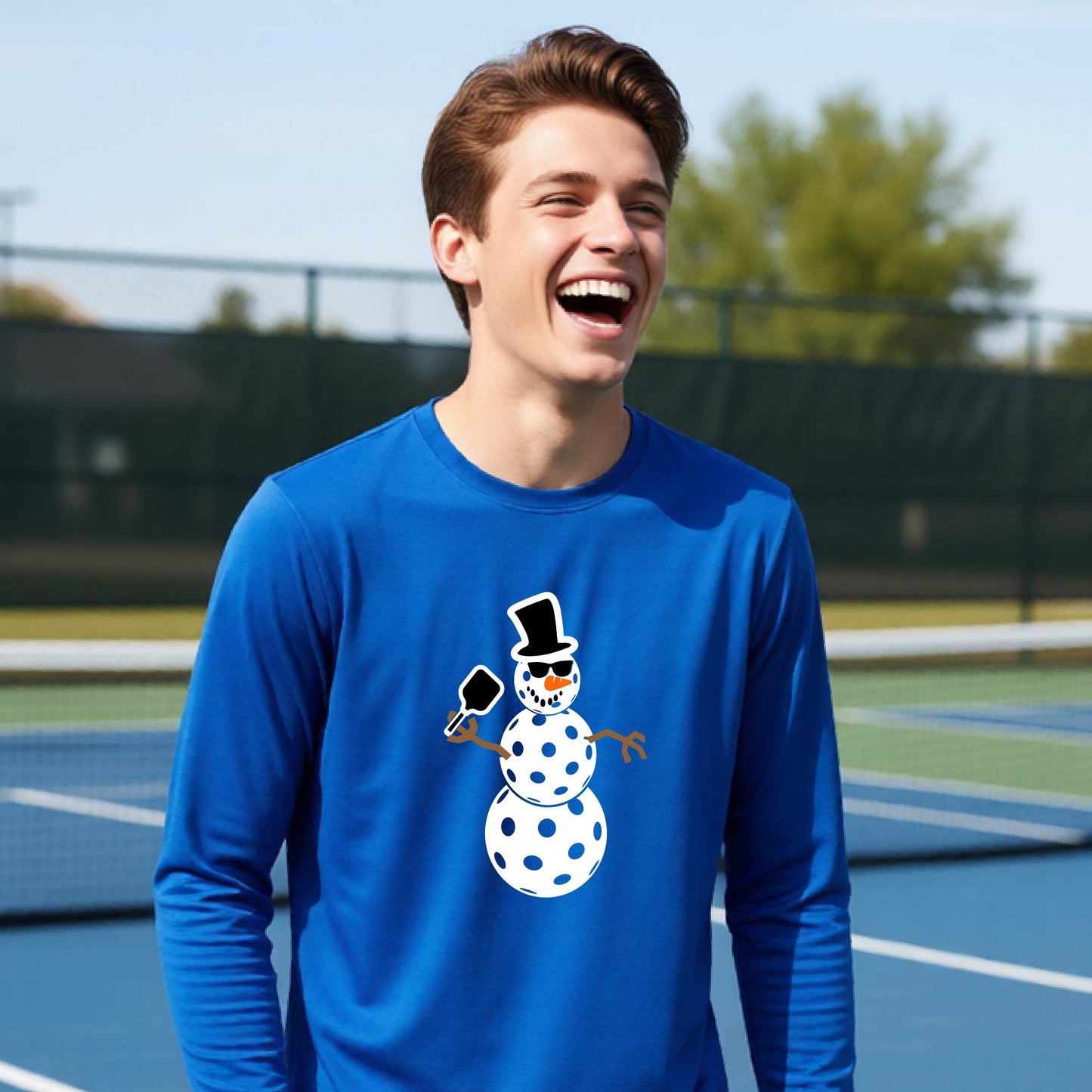A smiling young person stands on a pickleball court wearing a royal blue long-sleeve shirt with a white and pink polka-dot pickleball snowman graphic on the front.