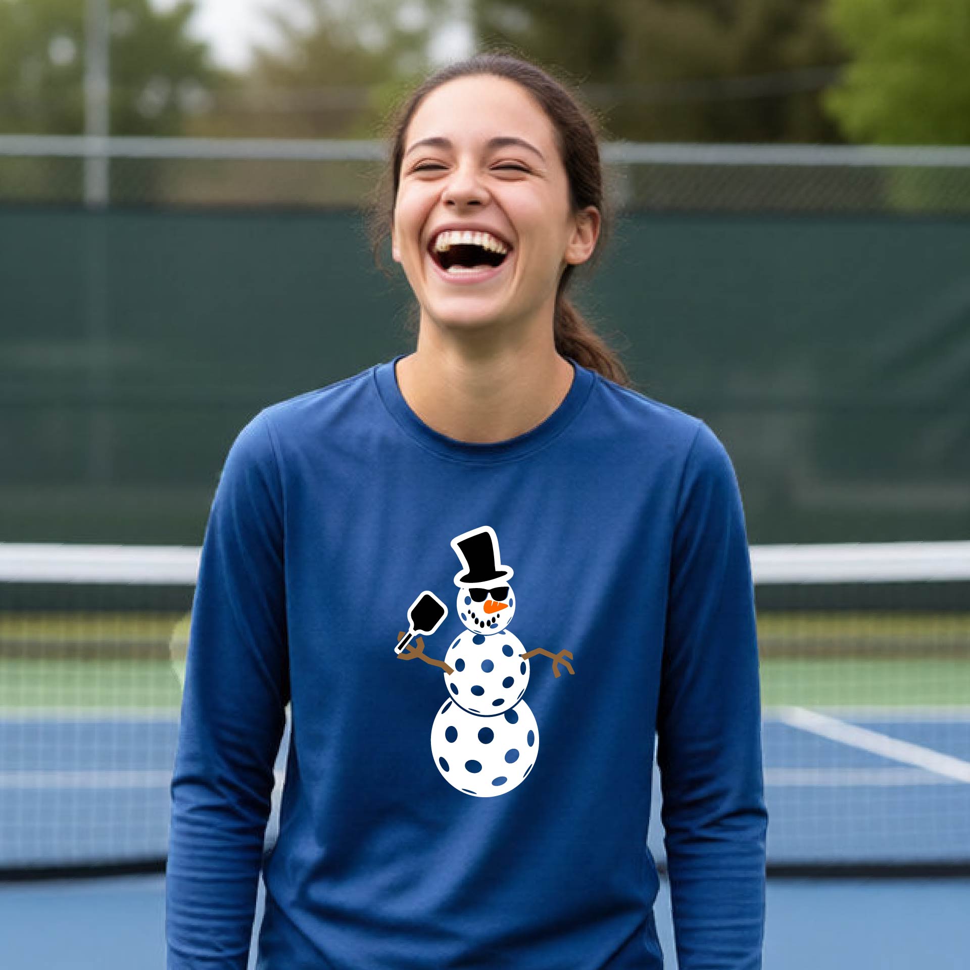 A smiling young person stands on a pickleball court wearing a royal blue long-sleeve shirt with a white and pink polka-dot pickleball snowman graphic on the front.