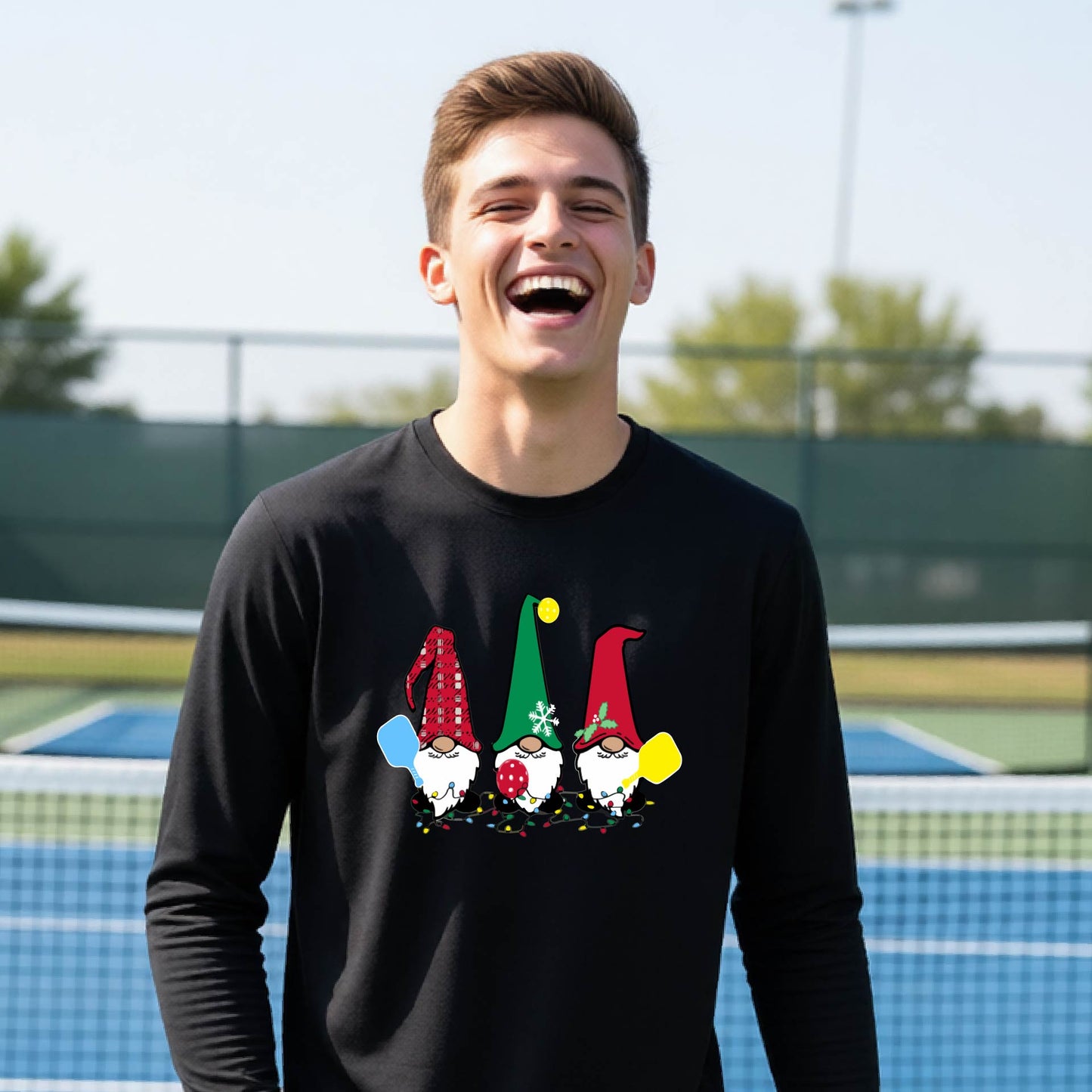 A young adult laughing on an outdoor pickleball  court wearing a black long-sleeve shirt with a graphic of three cartoon pickleball garden gnomes wearing christmas hats.