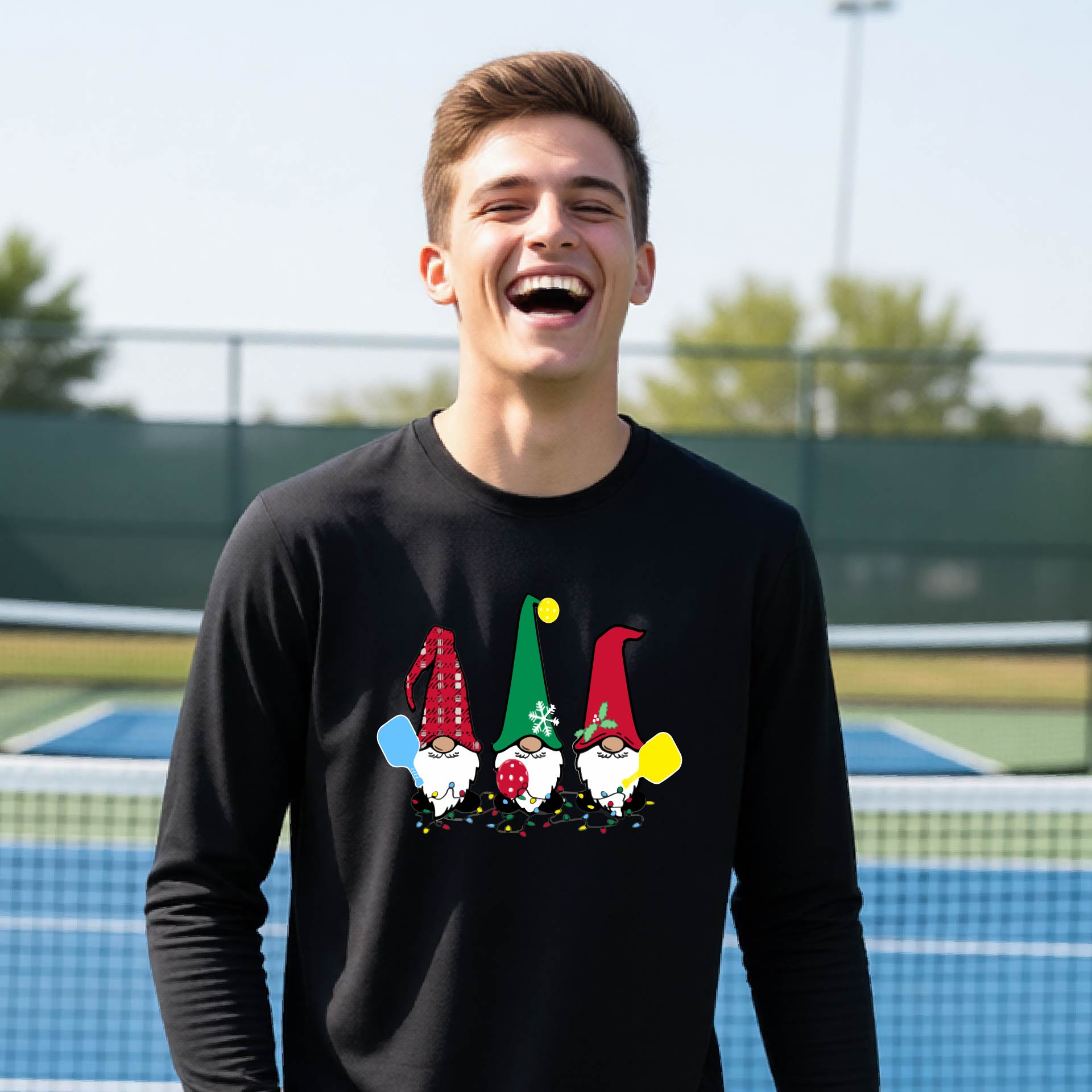 A young adult laughing on an outdoor pickleball  court wearing a black long-sleeve shirt with a graphic of three cartoon pickleball garden gnomes wearing christmas hats.