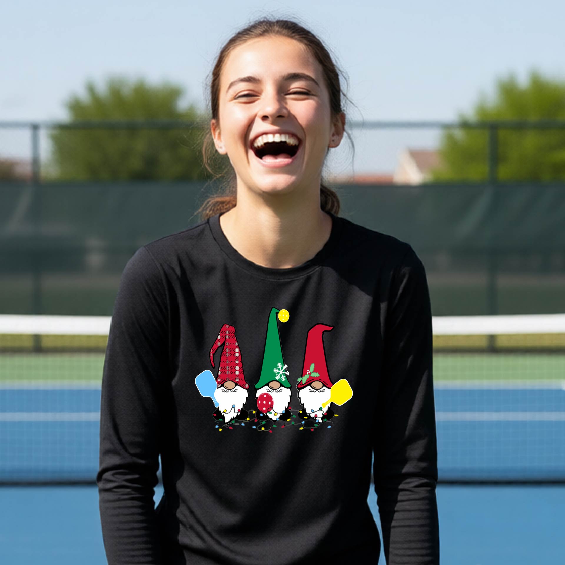 A young adult laughing on an outdoor pickleball  court wearing a black long-sleeve shirt with a graphic of three cartoon pickleball garden gnomes wearing christmas hats.