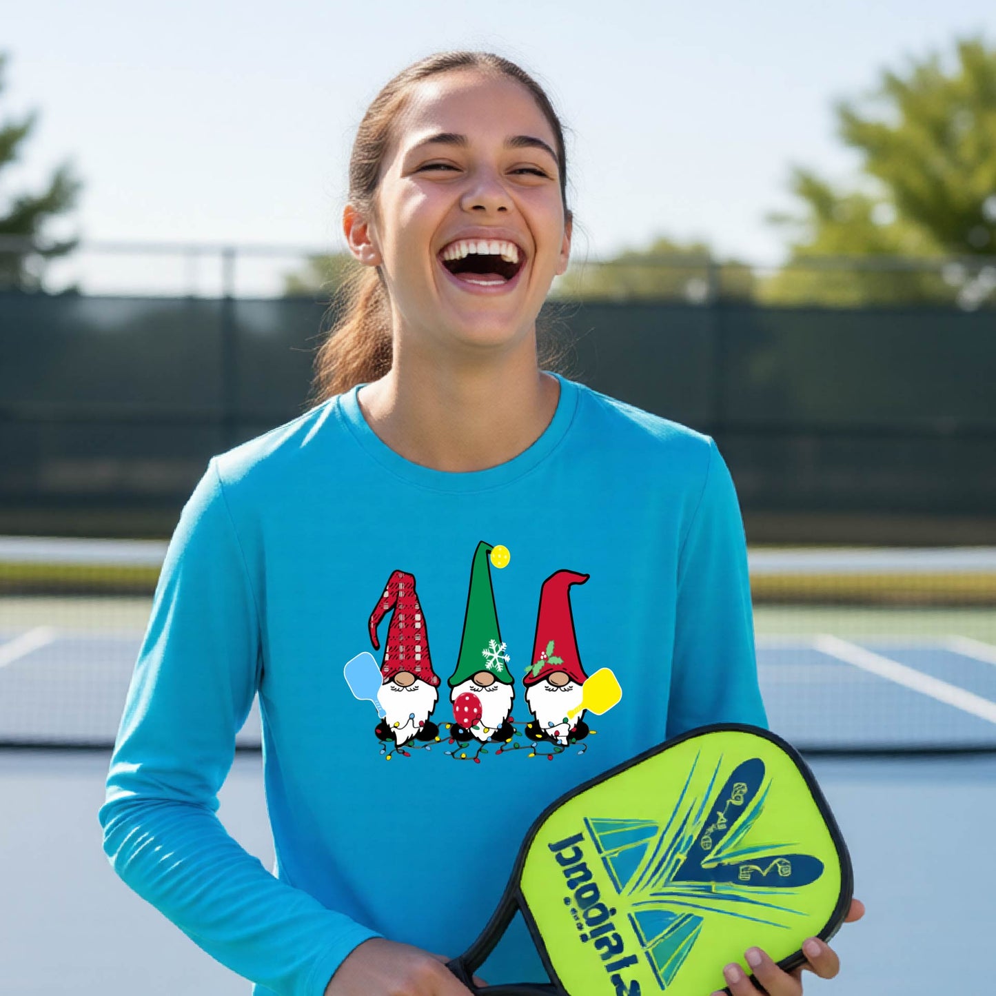 A young adult laughing on an outdoor pickleball  court wearing a bright blue long-sleeve shirt with a graphic of three cartoon pickleball garden gnomes wearing christmas hats.