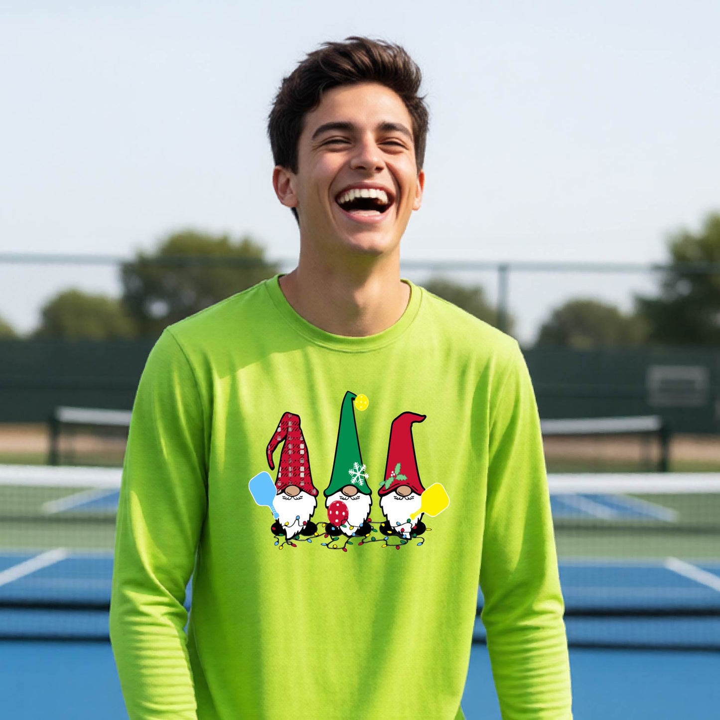 A young adult laughing on an outdoor pickleball  court wearing a bright lime green long-sleeve shirt with a graphic of three cartoon pickleball garden gnomes wearing christmas hats.