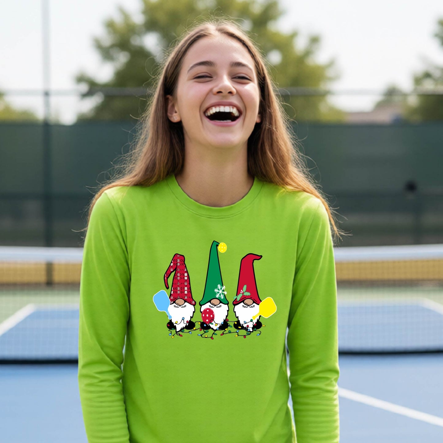 A young adult laughing on an outdoor pickleball  court wearing a lime green long-sleeve shirt with a graphic of three cartoon pickleball garden gnomes wearing christmas hats.
