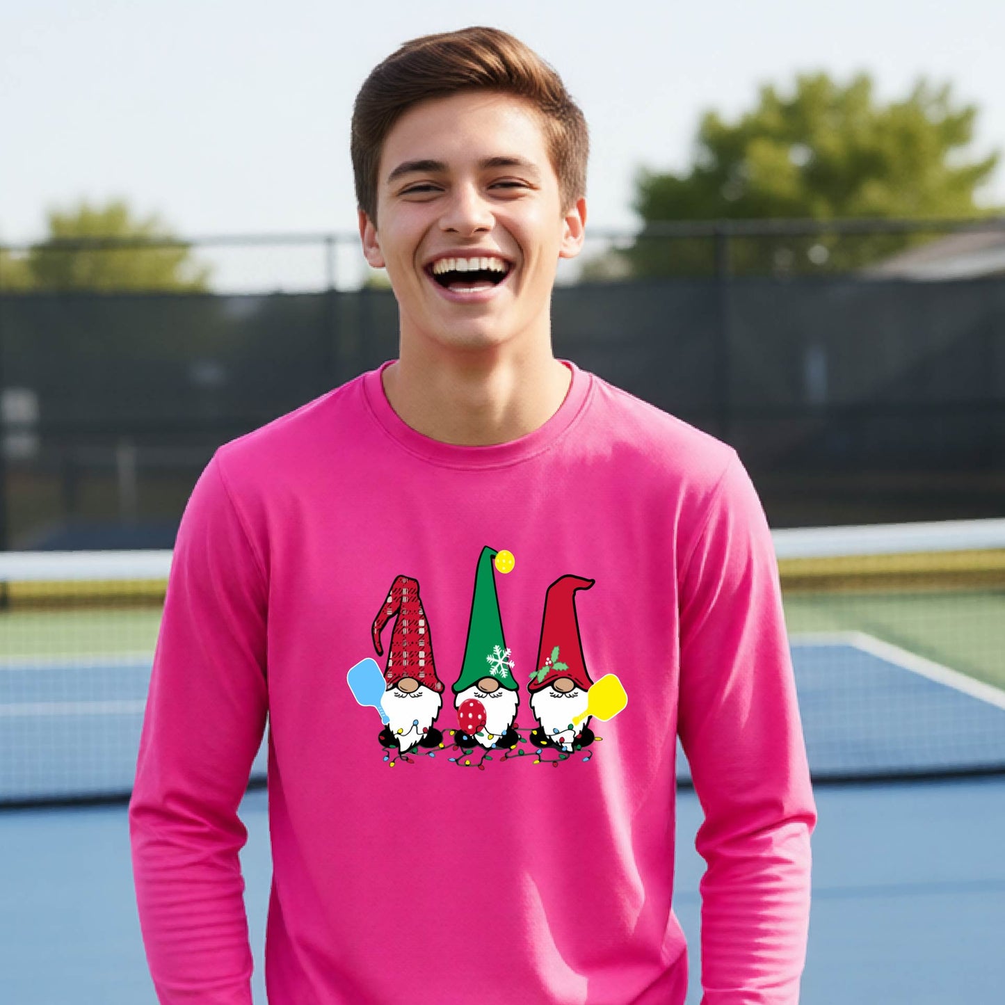 A young adult laughing on an outdoor pickleball  court wearing a bright pink long-sleeve shirt with a graphic of three cartoon pickleball garden gnomes wearing christmas hats.