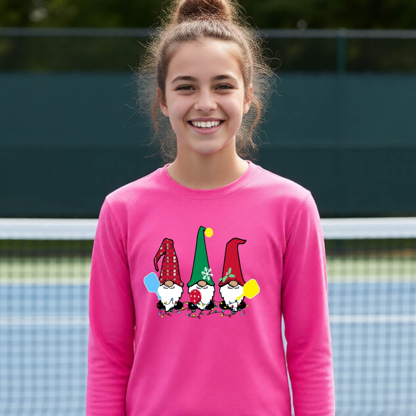 A young adult laughing on an outdoor pickleball  court wearing a bright pink long-sleeve shirt with a graphic of three cartoon pickleball garden gnomes wearing christmas hats.