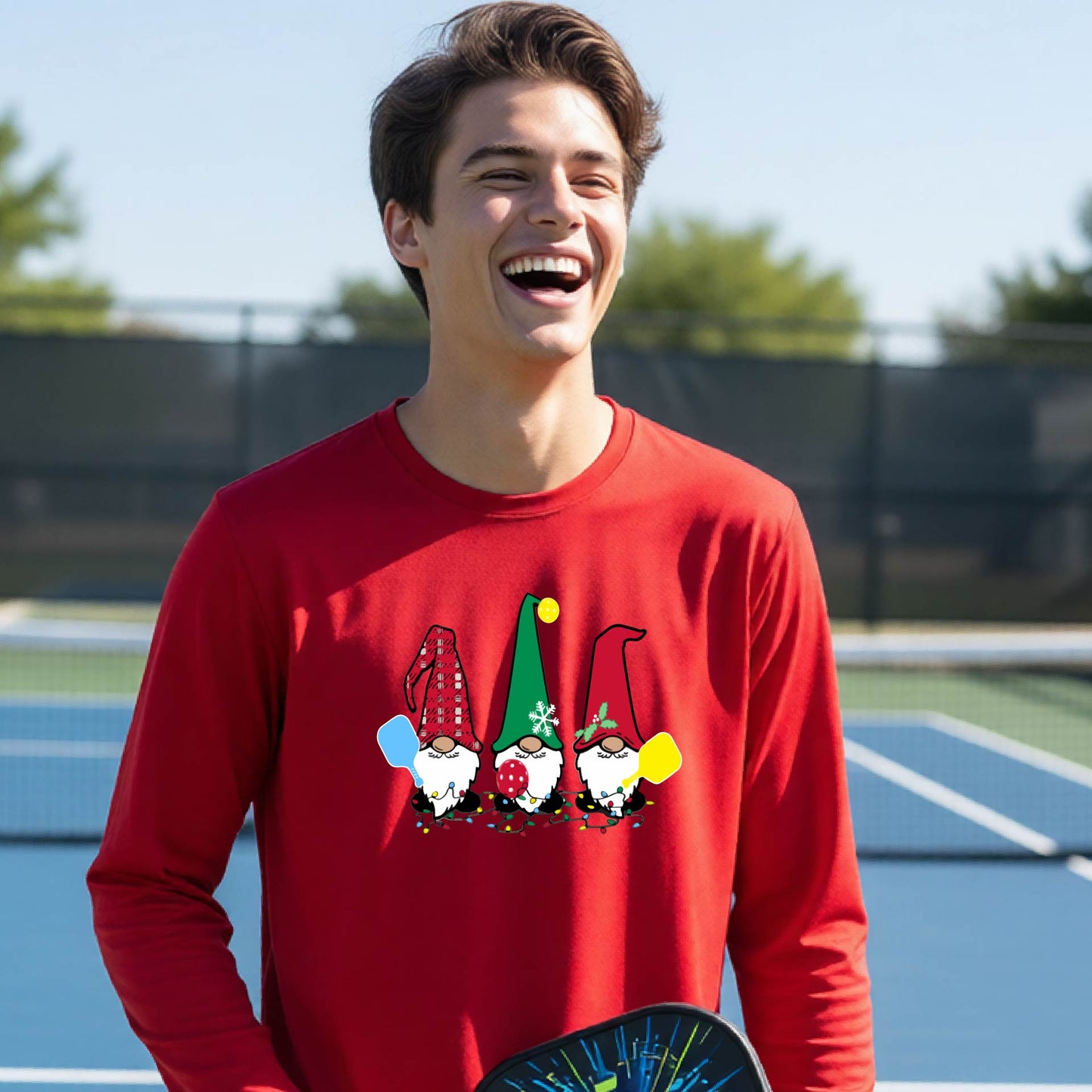 A young adult laughing on an outdoor pickleball  court wearing a red long-sleeve shirt with a graphic of three cartoon pickleball garden gnomes wearing christmas hats.