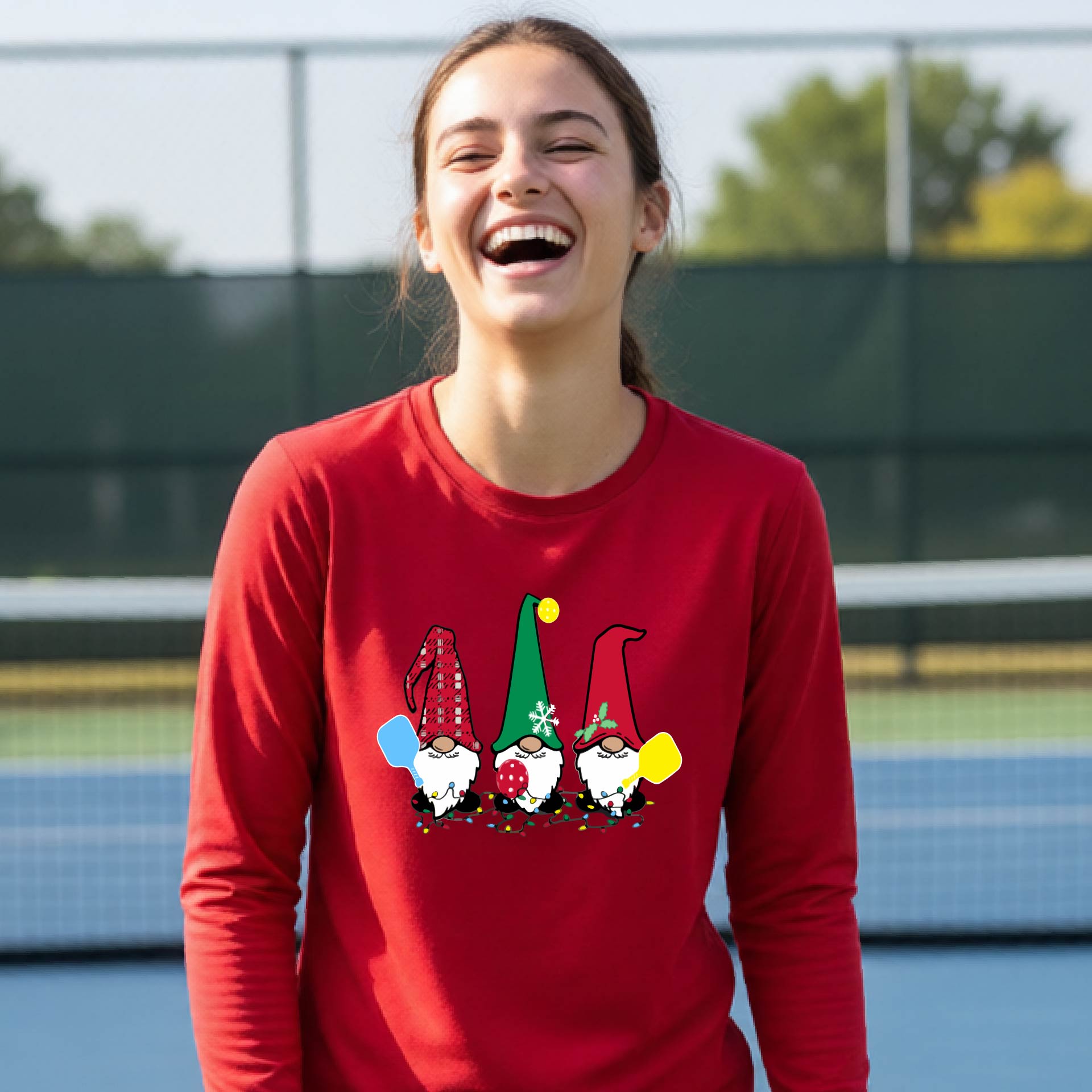 A young adult laughing on an outdoor pickleball  court wearing a red long-sleeve shirt with a graphic of three cartoon pickleball garden gnomes wearing christmas hats.gnomes.