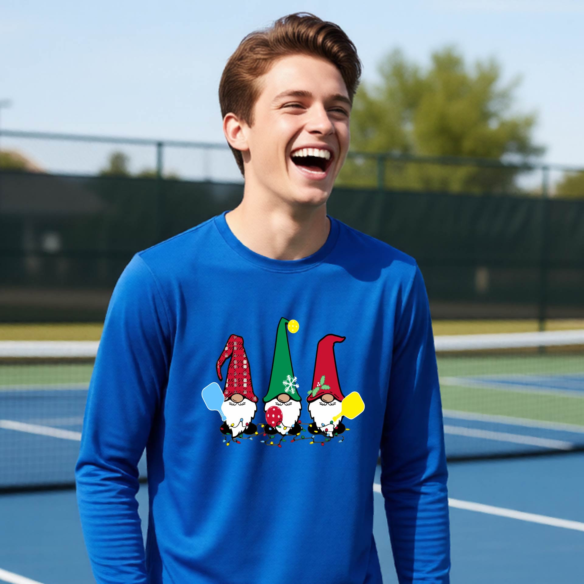 A young adult laughing on an outdoor pickleball  court wearing a royal blue long-sleeve shirt with a graphic of three cartoon pickleball garden gnomes wearing christmas hats.
