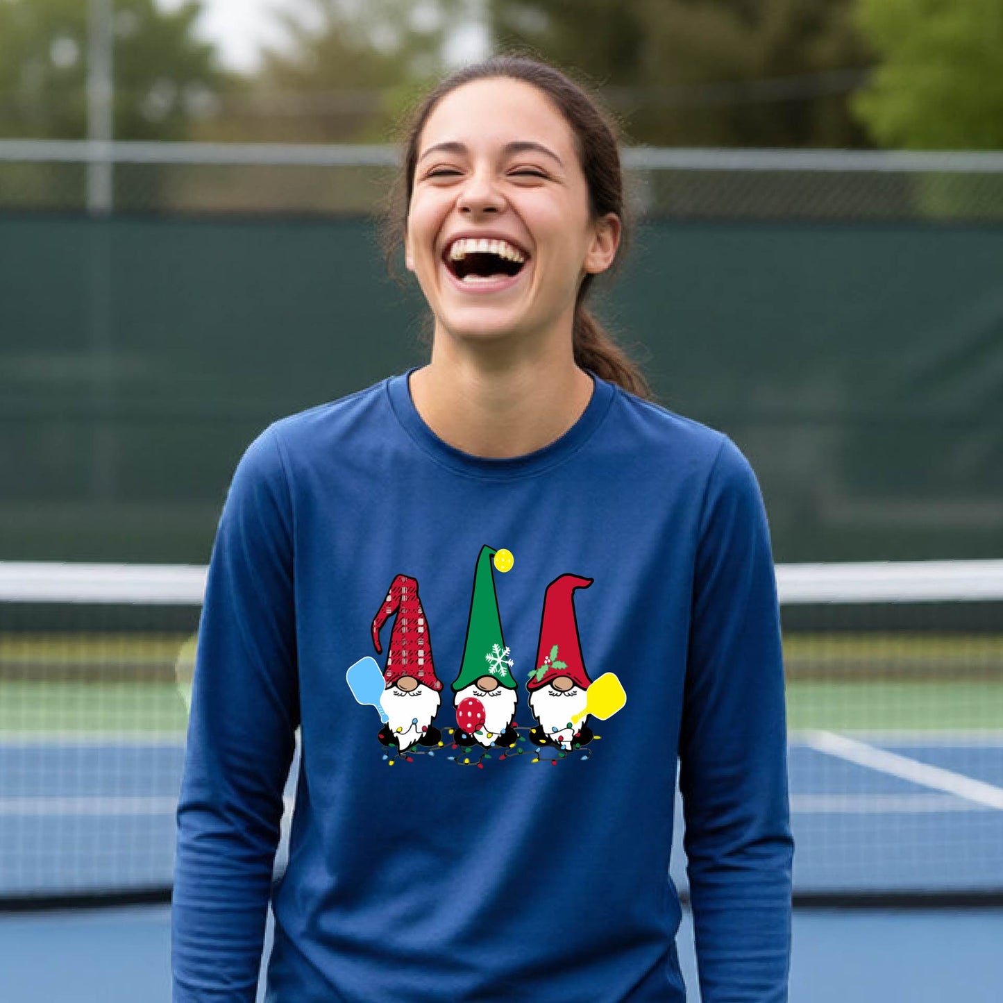 A young adult laughing on an outdoor pickleball  court wearing a royal blue long-sleeve shirt with a graphic of three cartoon pickleball garden gnomes wearing christmas hats.