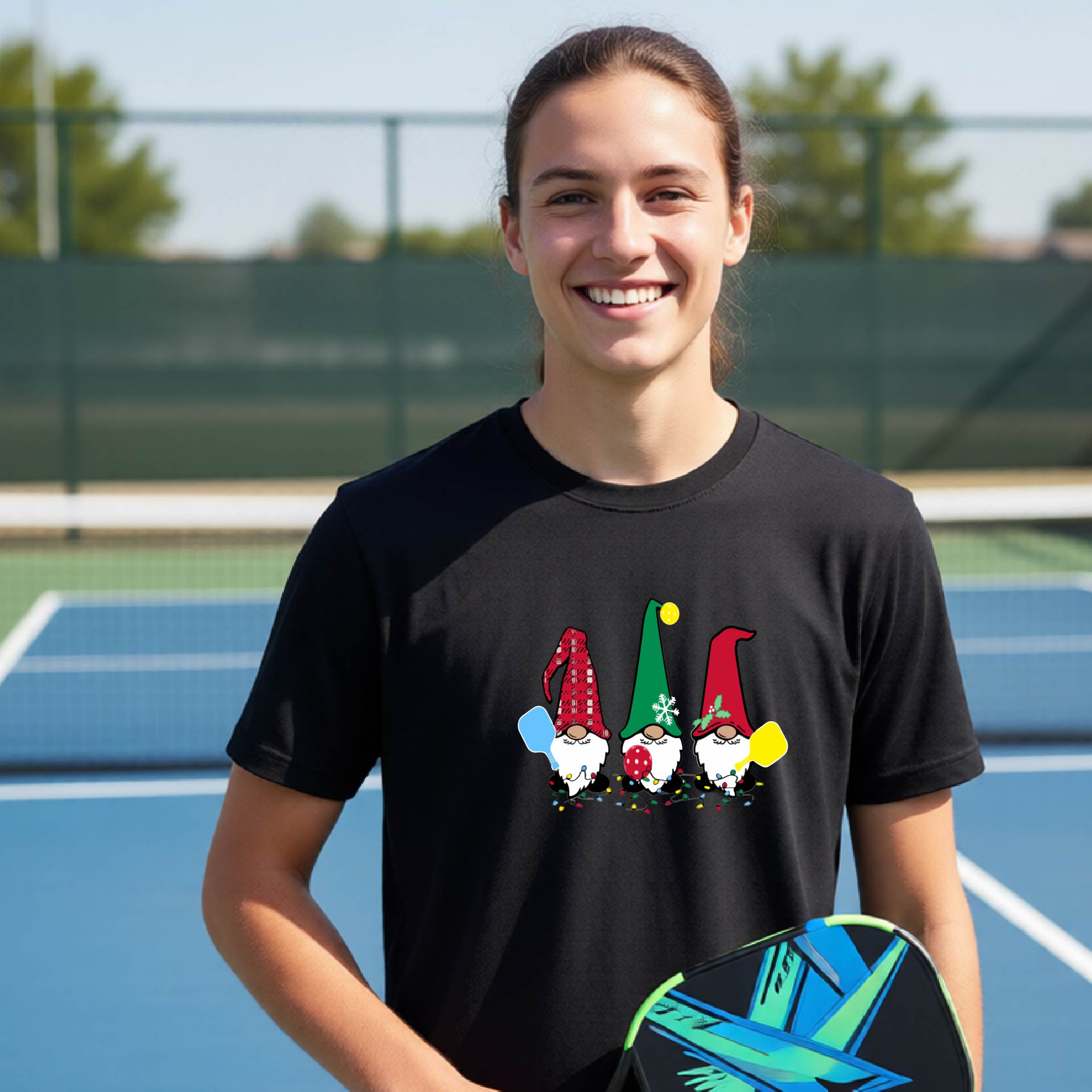 A smiling young person on a blue pickleball court wearing a black t-shirt with a colorful gnome graphic on the chest, holding a pickleball paddle.