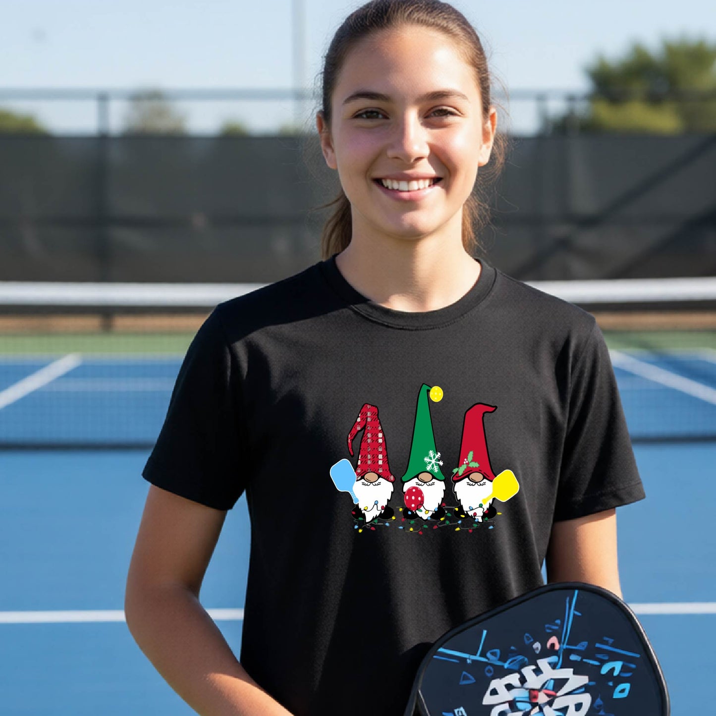 A smiling young person on a blue pickleball court wearing a black t-shirt with a colorful gnome graphic on the chest, holding a pickleball paddle.