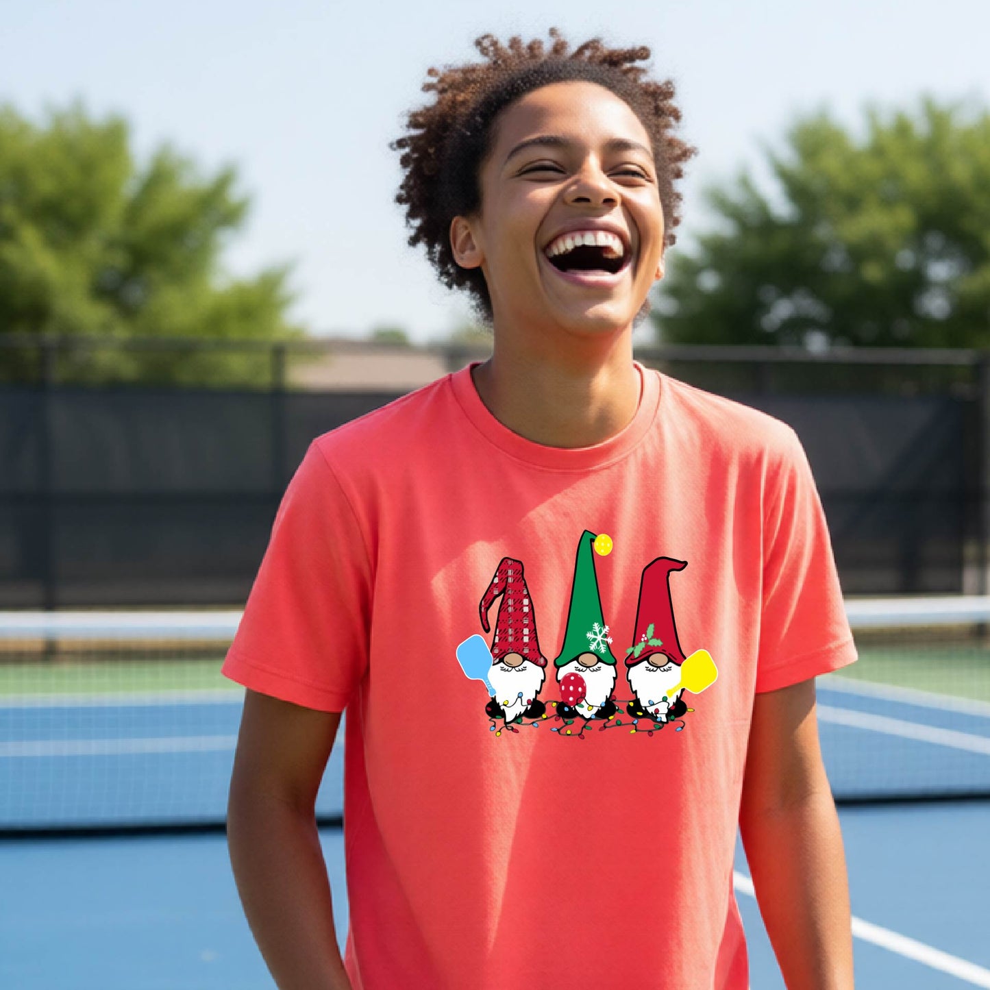A woman laughing on an outdoor pickleball court wearing a coral-colored crewneck t-shirt with a graphic of three festive garden pickleball gnomes.