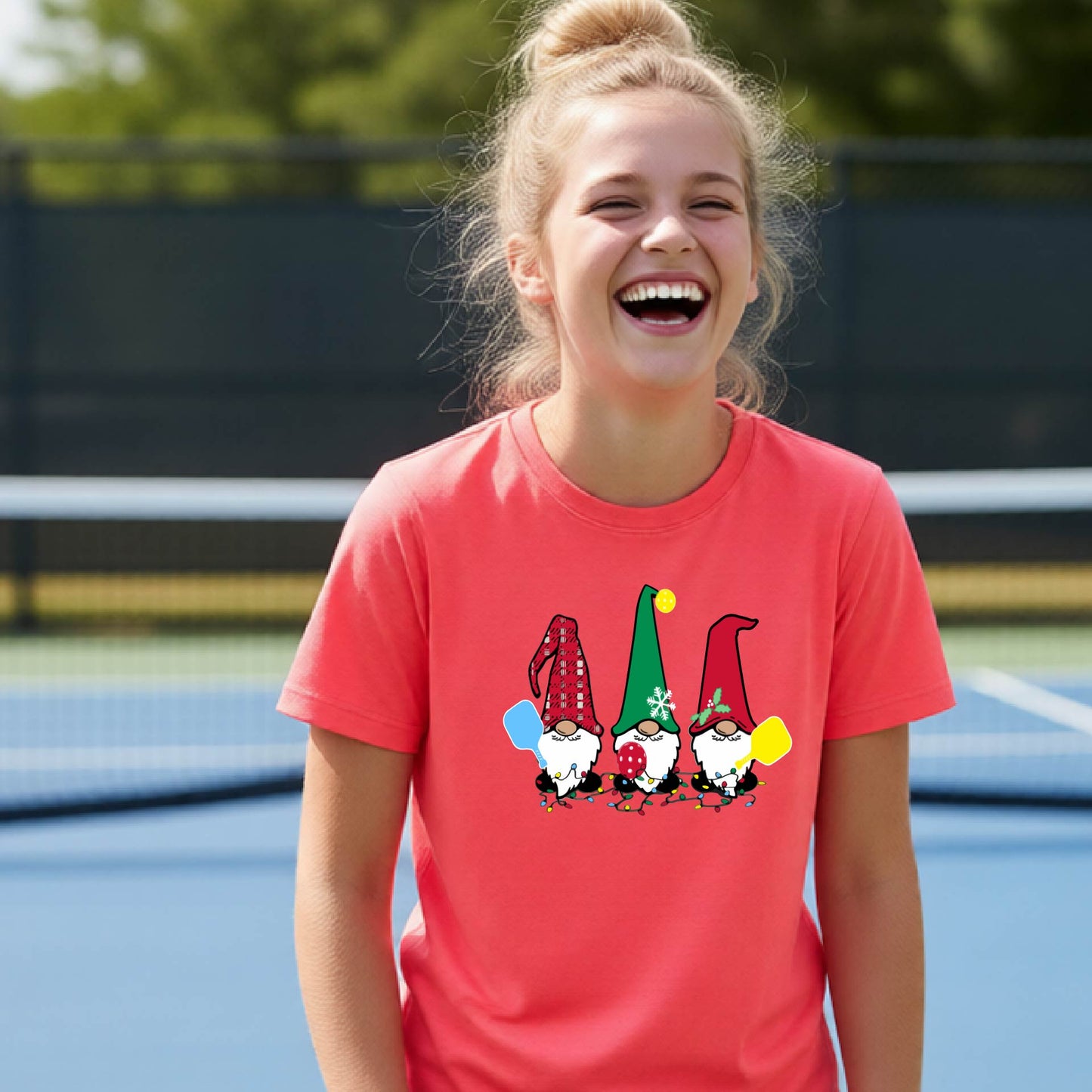 A smiling young person on a blue pickleball court wearing a coral-colored t-shirt with a colorful gnome graphic on the chest, holding a pickleball paddle.