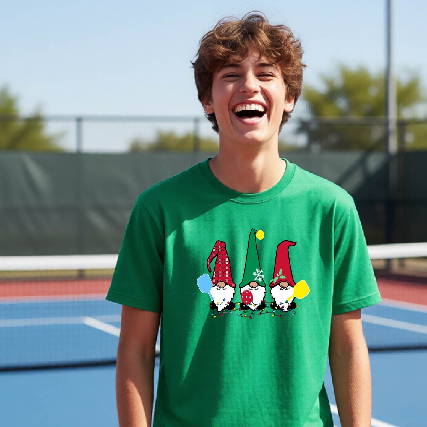 A smiling young person on a blue pickleball court wearing a green t-shirt with a colorful gnome graphic on the chest, holding a pickleball paddle.