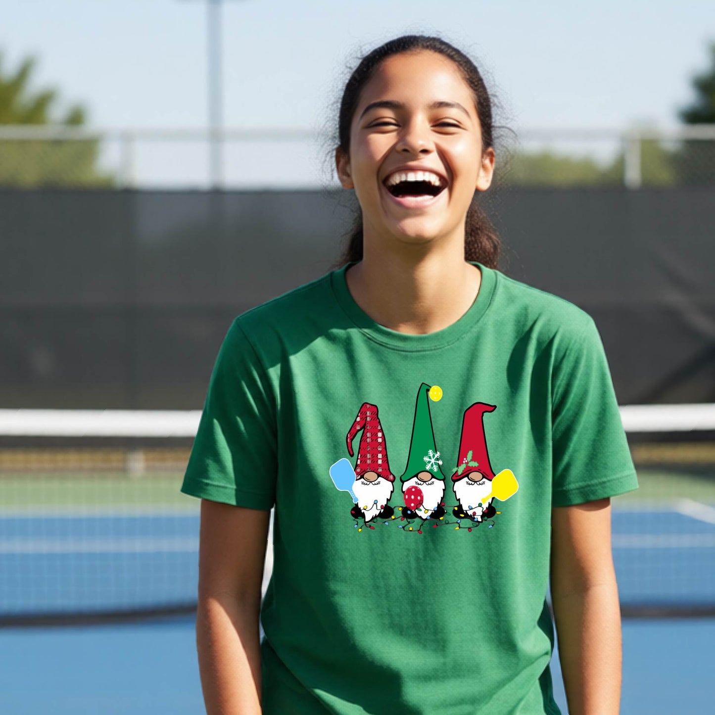 A woman laughing on an outdoor pickleball court wearing a green crewneck t-shirt with a graphic of three festive garden pickleball gnomes.