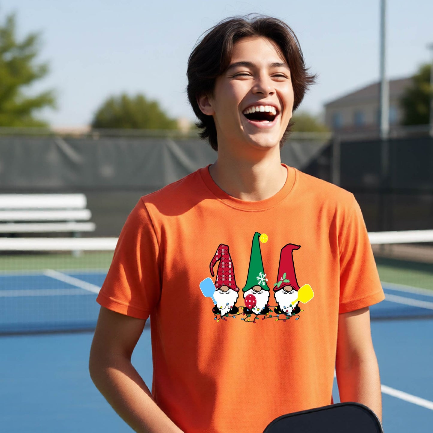 A woman laughing on an outdoor pickleball court wearing an orange crewneck t-shirt with a graphic of three festive garden pickleball gnomes.
