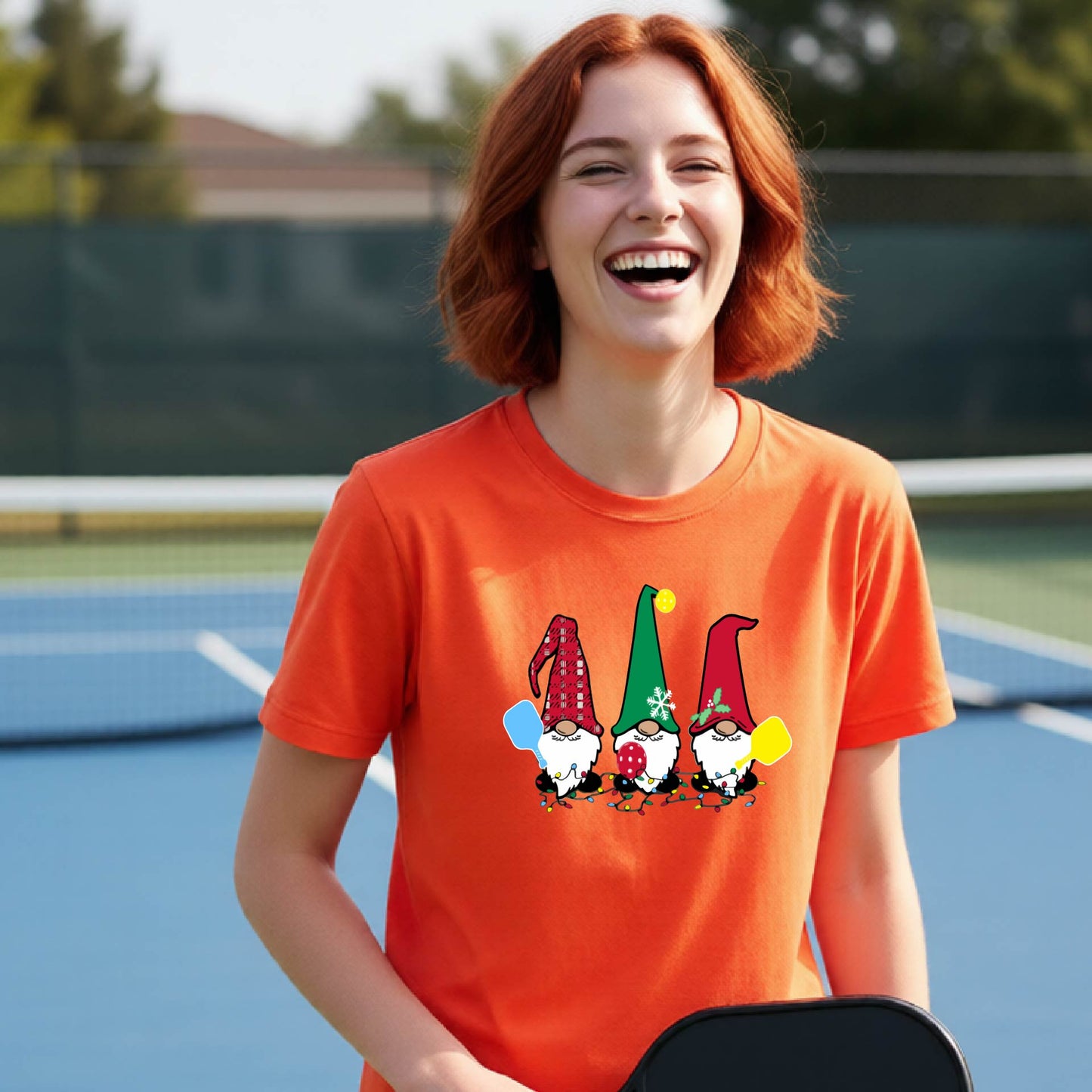 A smiling young person on a blue pickleball court wearing an orange t-shirt with a colorful gnome graphic on the chest, holding a pickleball paddle.