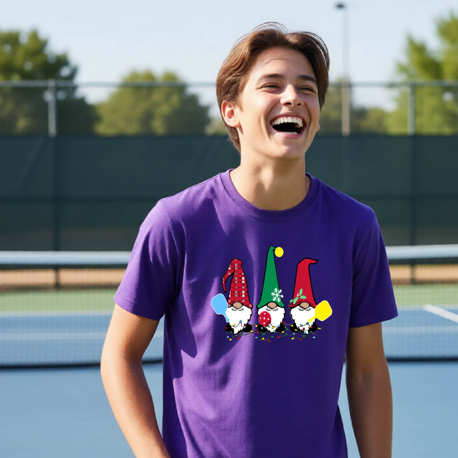 A smiling young person on a blue pickleball court wearing a purple t-shirt with a colorful gnome graphic on the chest, holding a pickleball paddle.