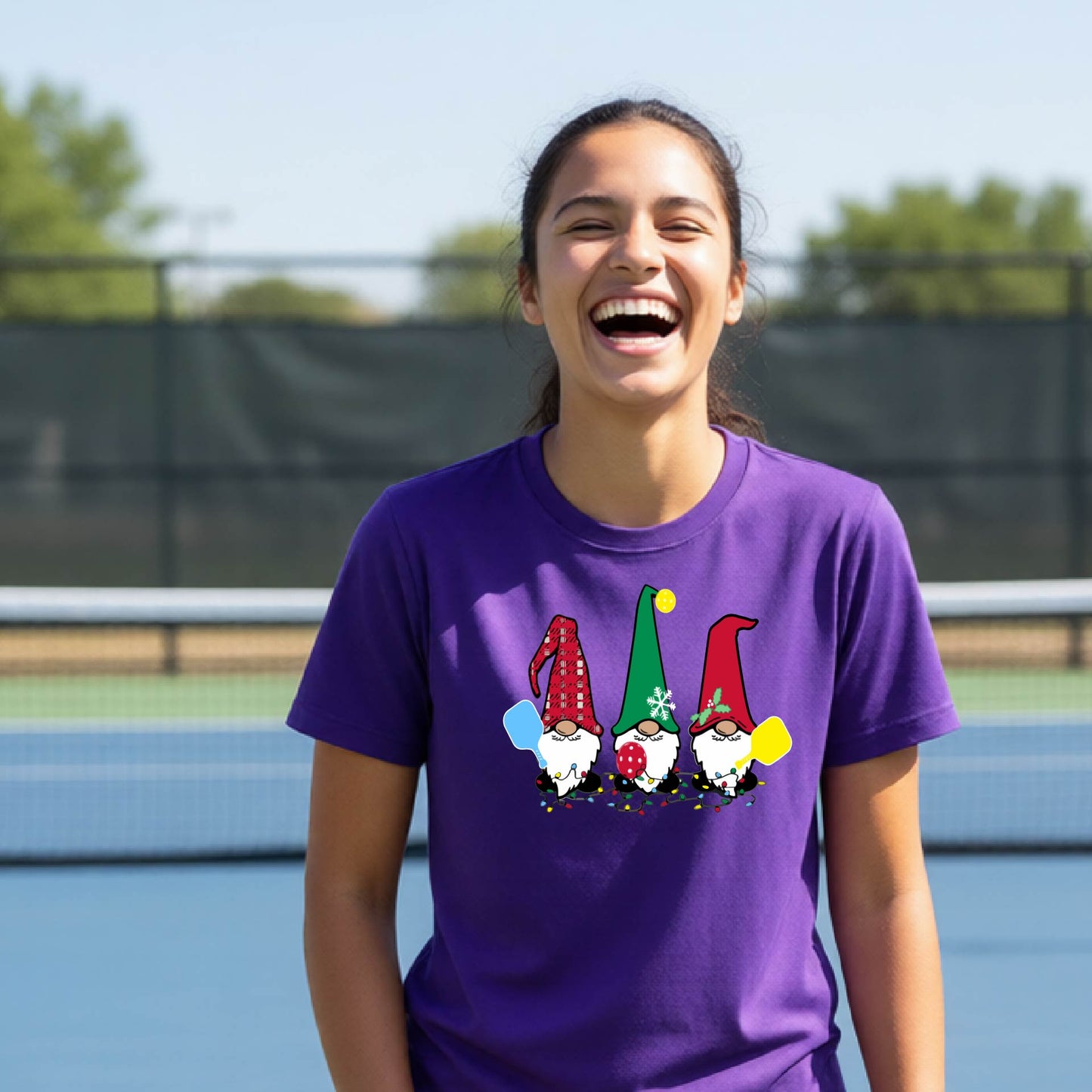 A woman laughing on an outdoor pickleball court wearing a purple crewneck t-shirt with a graphic of three festive garden pickleball gnomes.