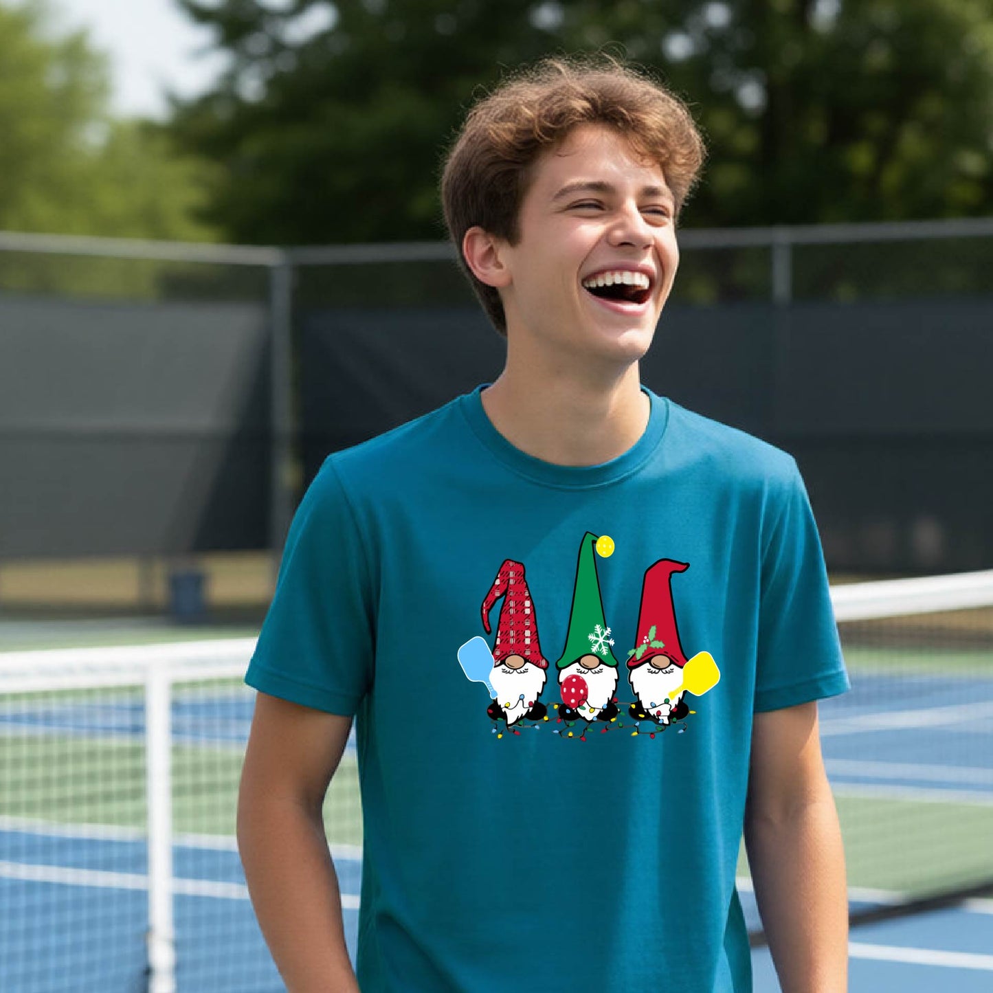 A smiling young person on a blue pickleball court wearing a teal t-shirt with a colorful gnome graphic on the chest, holding a pickleball paddle.