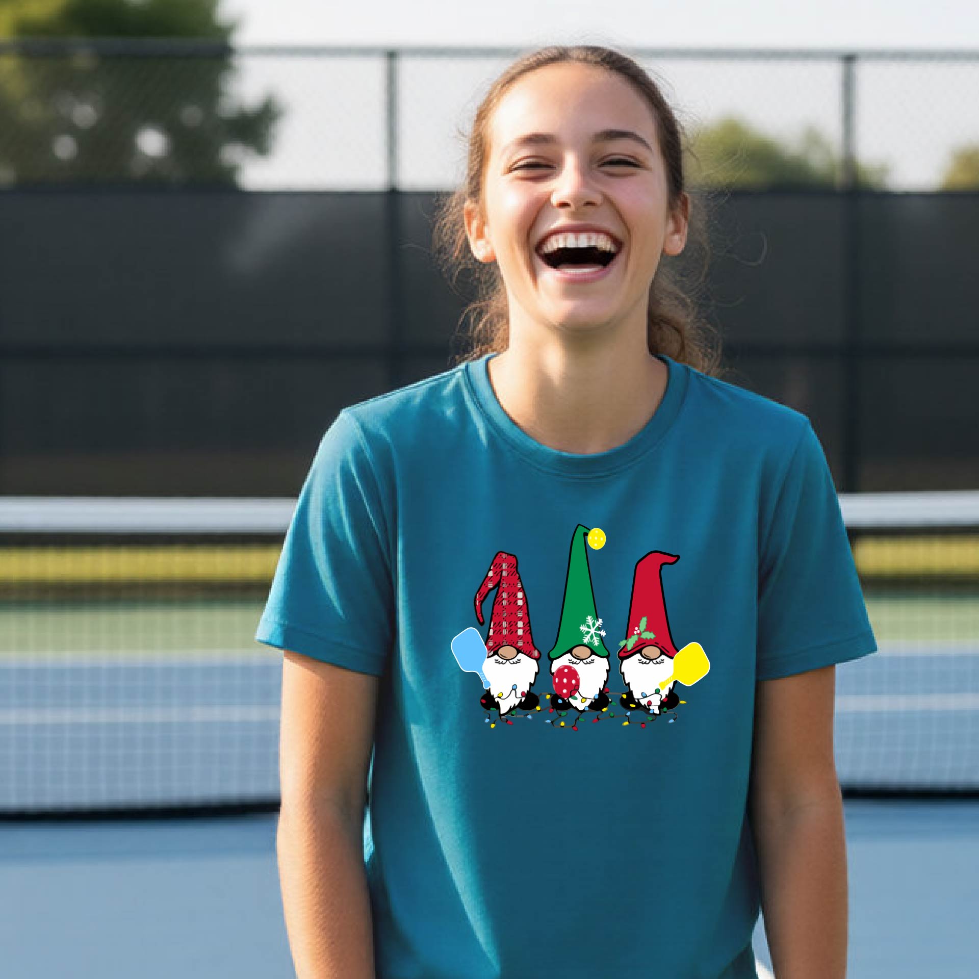 A woman laughing on an outdoor pickleball court wearing a teal crewneck t-shirt with a graphic of three festive garden pickleball gnomes.
