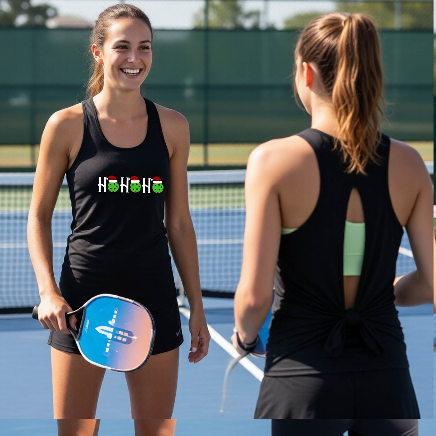 Two women on an outdoor pickleball court wearing matching black split-back athletic tank tops. One woman faces forward, showing a front graphic with a green pickleball wearing white/red Santa hats. 