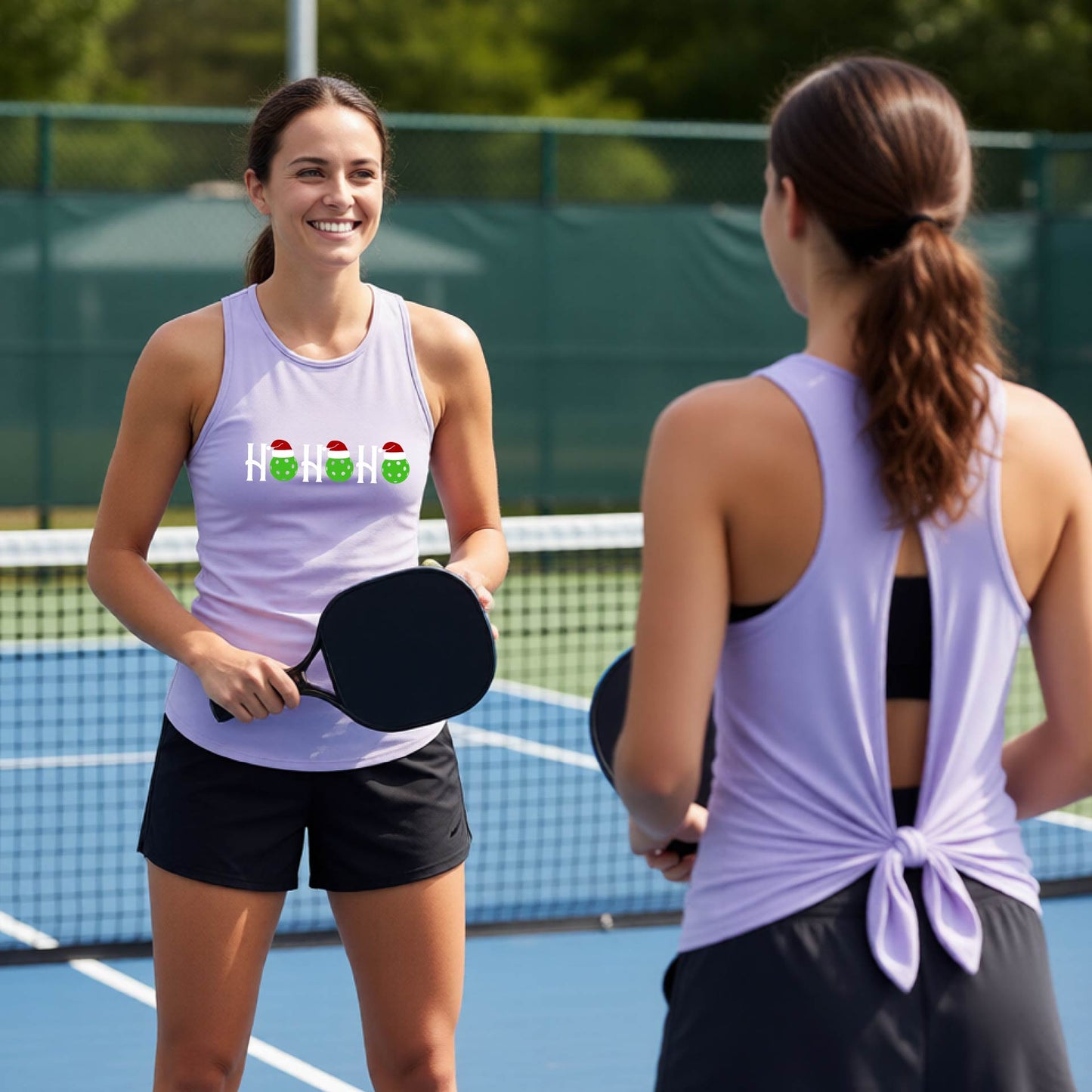 Two women on an outdoor pickleball court wearing matching light lavender purple split-back athletic tank tops. One woman faces forward, showing a front graphic with a green pickleball wearing white/red Santa hats.