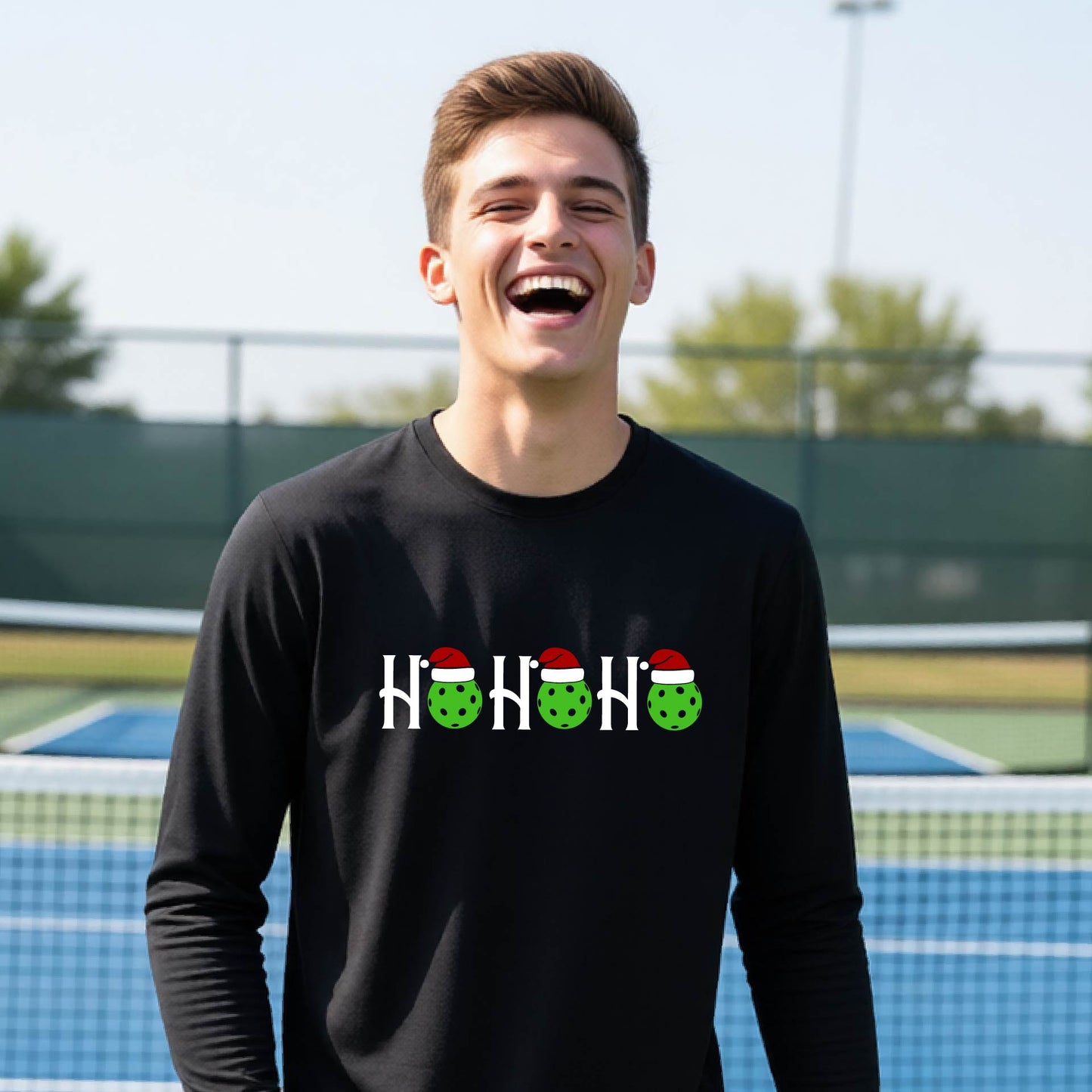 A smiling young person stands on pickleball court wearing a black long-sleeve shirt with a 'HO HO HO' graphic made of green pickleball ornaments wearing red Santa hats.