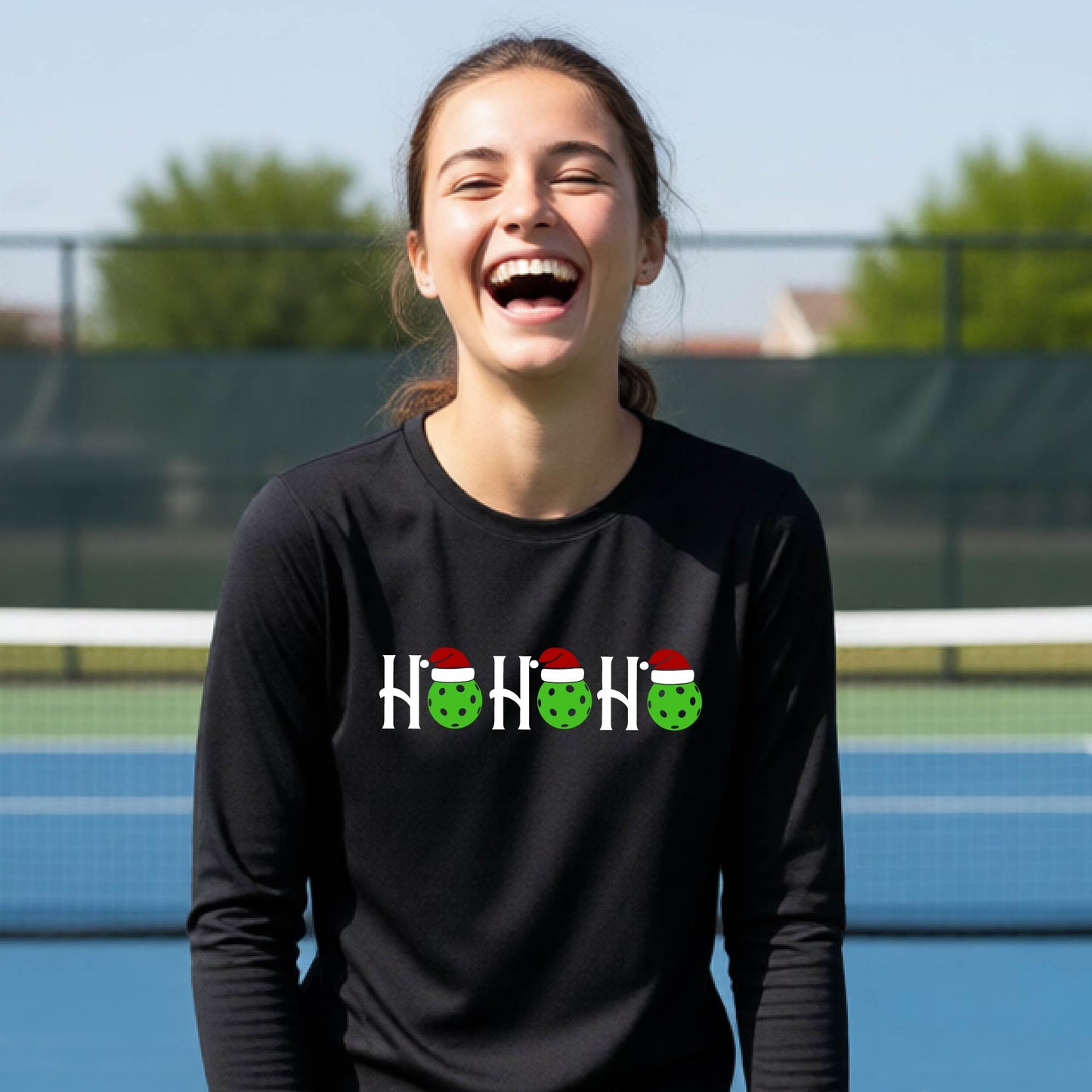 A smiling young person stands on pickleball court wearing a black long-sleeve shirt with a 'HO HO HO' graphic made of green pickleball ornaments wearing red Santa hats.