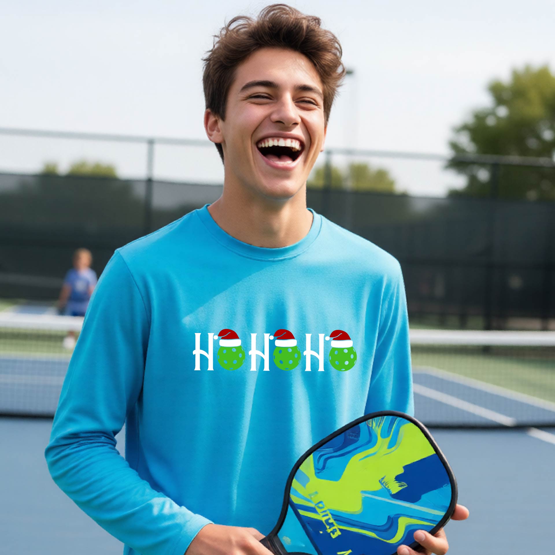 A smiling young person stands on pickleball court wearing a bright blue long-sleeve shirt with a 'HO HO HO' graphic made of green pickleball ornaments wearing red Santa hats.