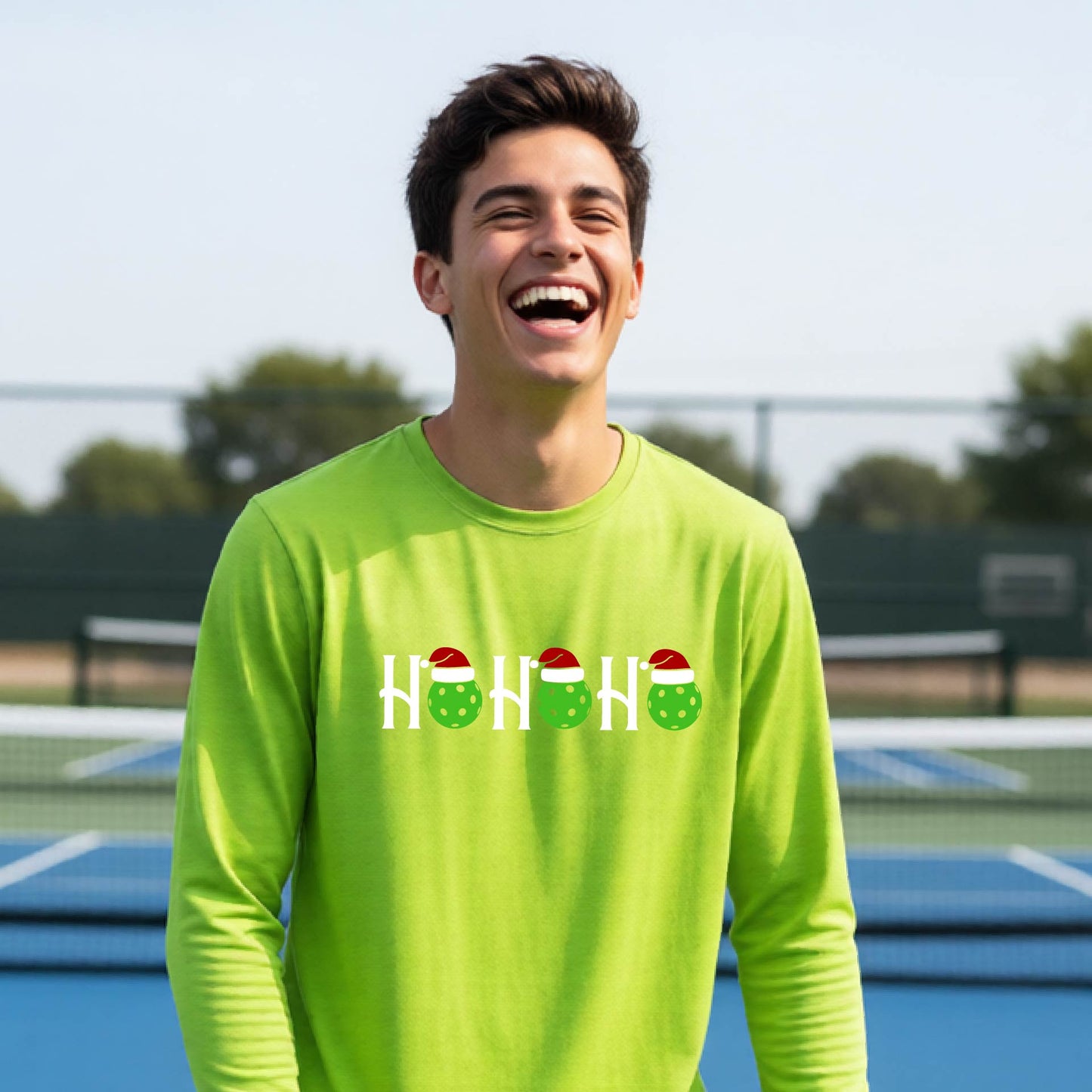 A smiling young person stands on pickleball court wearing a bright lime green long-sleeve shirt with a 'HO HO HO' graphic made of green pickleball ornaments wearing red Santa hats.