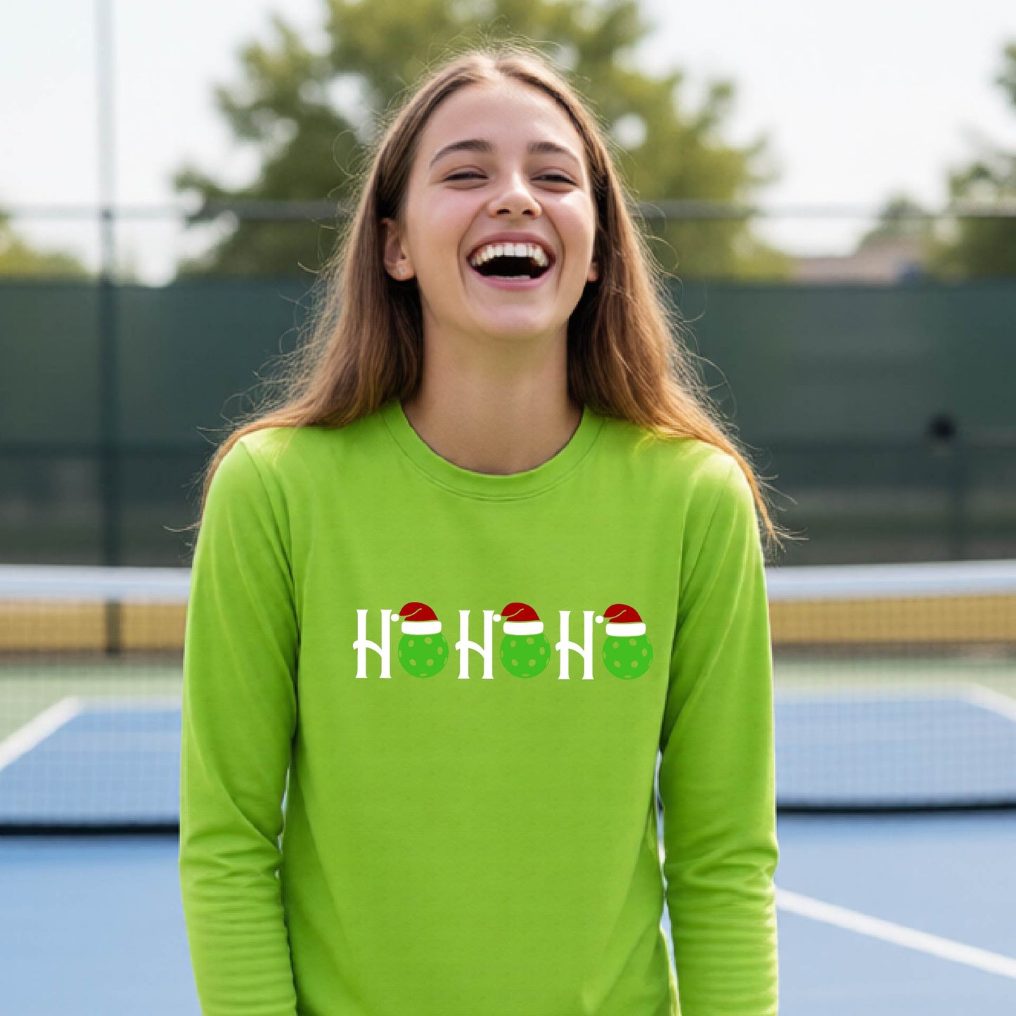 A smiling young person stands on pickleball court wearing a bright lime green long-sleeve shirt with a 'HO HO HO' graphic made of green pickleball ornaments wearing red Santa hats.