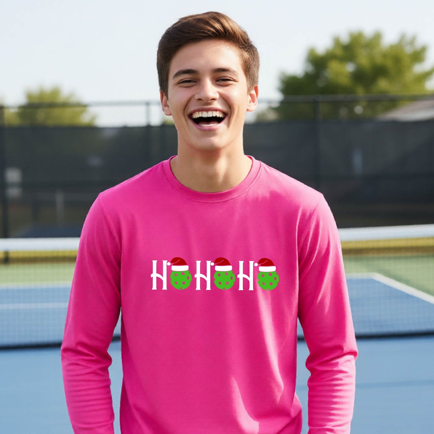 A smiling young person stands on pickleball court wearing a bright pink long-sleeve shirt with a 'HO HO HO' graphic made of green pickleball ornaments wearing red Santa hats.
