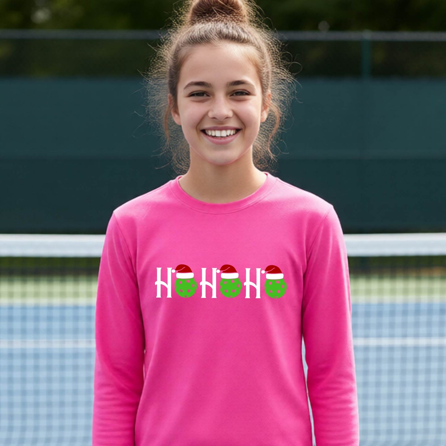 A smiling young person stands on pickleball court wearing a bright pink long-sleeve shirt with a 'HO HO HO' graphic made of green pickleball ornaments wearing red Santa hats.