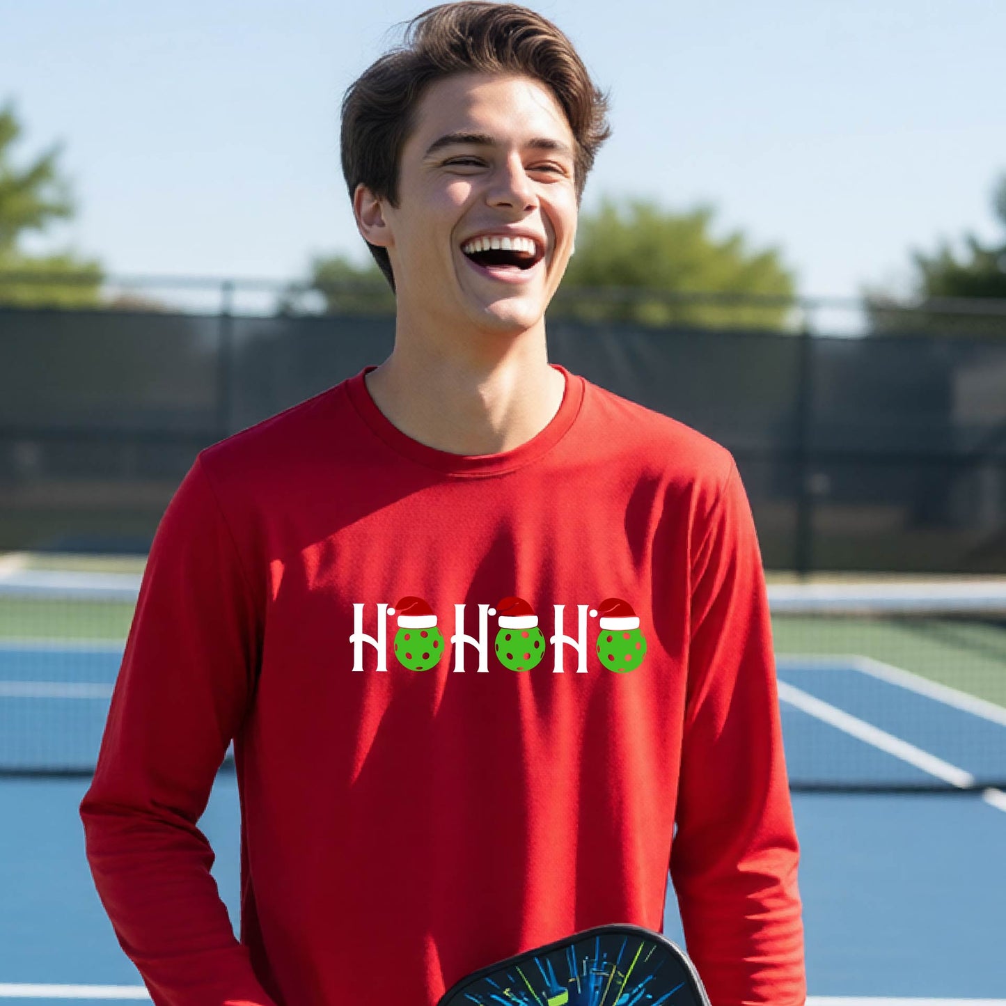 A smiling young person stands on pickleball court wearing a bright red long-sleeve shirt with a 'HO HO HO' graphic made of green pickleball ornaments wearing red Santa hats.